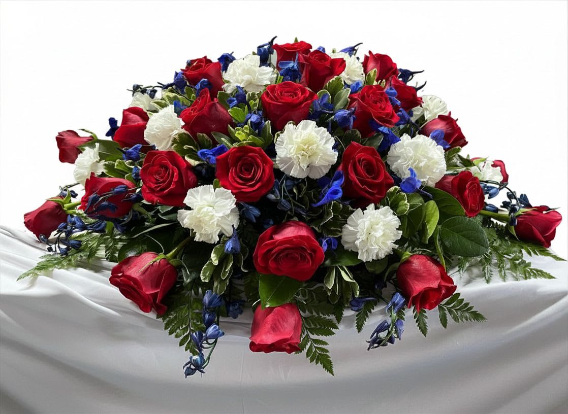 Liberty's Grace - A bold red, white and blue centerpiece designed to make someone feel honored and remembered, perfect as a patriotic table arrangement or memorial tribute. This flower arrangement for remembrance and ceremonies places red roses, white carnations and small blue accent blooms in a low, rounded composition so the recipient or honored guest looks straight into a lush carpet of flowers.  The design is dense and formal: tightly layered roses and carnations create a full surface with blooms spilling slightly over the edges for a generous, sculpted silhouette. Small blue flowers punctuate the palette and fresh green foliage lifts the colors; the arrangement sits on a draped white tablecloth for a ceremonial presentation that reads as a large centerpiece or tribute rather than a casual bouquet.  Choose this piece when you want the person you're honoring to feel truly seen-whether for a memorial service, Veterans Day display, or patriotic event. The low profile works well on tabletops and altars; please contact us for size options or delivery and we'll ensure the arrangement arrives ready to display on its white drape.  As similar as possible. Designer may make appropriate substitutions based on flower availability. 