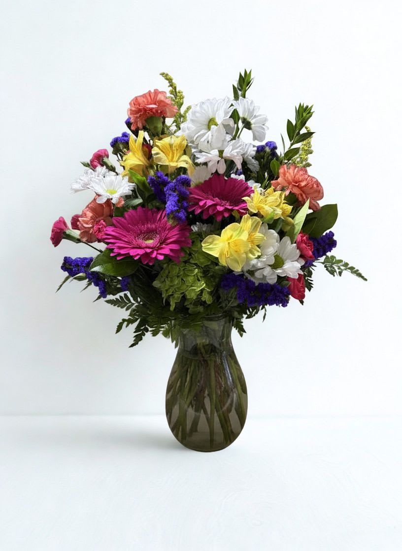 Colorful Oasis  - Bright pink gerbera daisies take center stage in this Colorful Oasis birthday bouquet, surrounded by cheerful white daisies, sunny yellow blooms, coral carnations, and pops of purple filler for contrast. Designed for birthdays, thank-you gestures, or spring celebrations, this rounded vase arrangement reads as upbeat and celebratory-the kind of gift that makes the person you're thinking of feel noticed and joyfully celebrated.  The full, garden-style composition is arranged in a clear glass vase that showcases the gathered stems for a fresh-from-the-garden look. Textures range from the bold, flat faces of gerberas to the ruffled carnations and delicate daisy petals, creating lively movement and visual depth in pinks, whites, yellows, corals, and purples. Size and stem length are balanced to present a dense, rounded silhouette that fits well on a table, desk, or mantel.  Perfect for sending warm wishes to a friend, brightening a coworker's day, or marking a springtime moment, this arrangement arrives ready to display-just add fresh water and recut stems as needed to extend blooms. The overall effect is vibrant and sincere: a colorful oasis that leaves the recipient smiling.