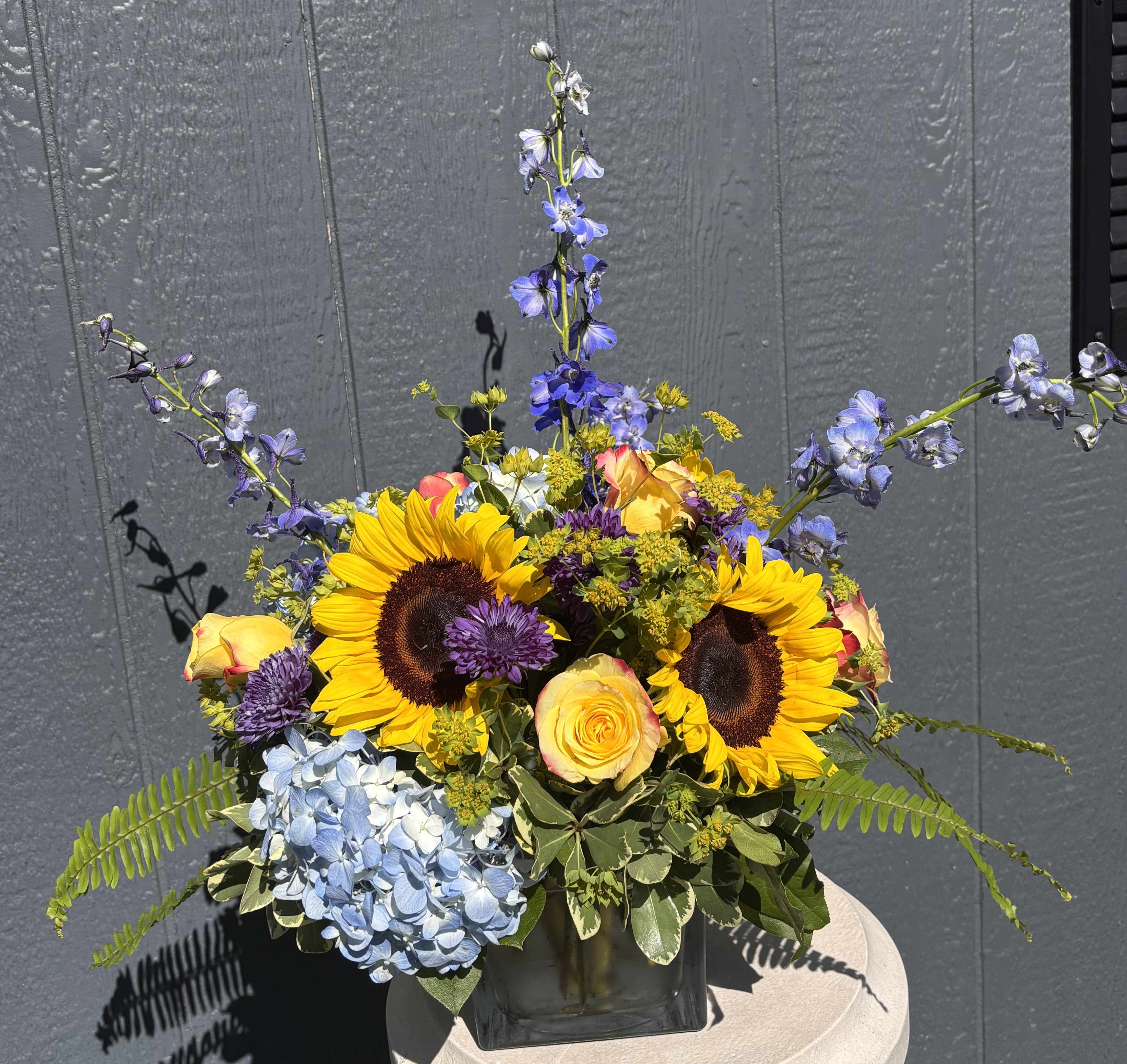 Sunflower Meadow - Bring the beauty of a perfect summer day indoors with our Sunflower Meadow arrangement. Featuring vibrant sunflowers, soft blue hydrangea, striking blue delphinium, sunny roses, blue button mums, and airy bupleurum, this design captures the warmth of sunshine beneath a bright blue sky.  Arranged in a modern glass cube, this cheerful arrangement is perfect for birthdays, celebrations, thank-you gifts, or simply brightening someone's day.