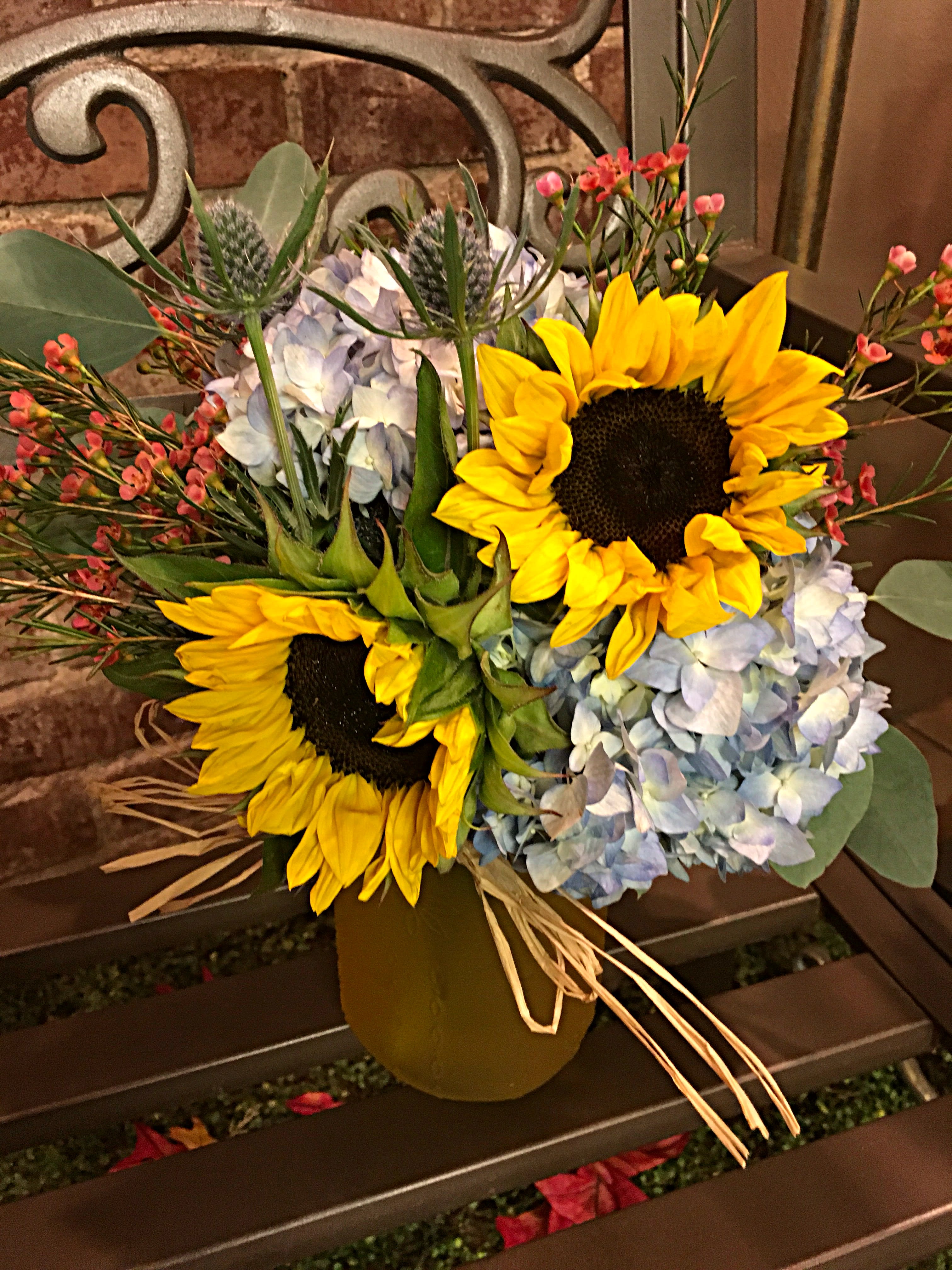 Harvest Moon - Blue hydrangea, thistle, sunflowers and seasonal accent flowers arranged in a colored vase.