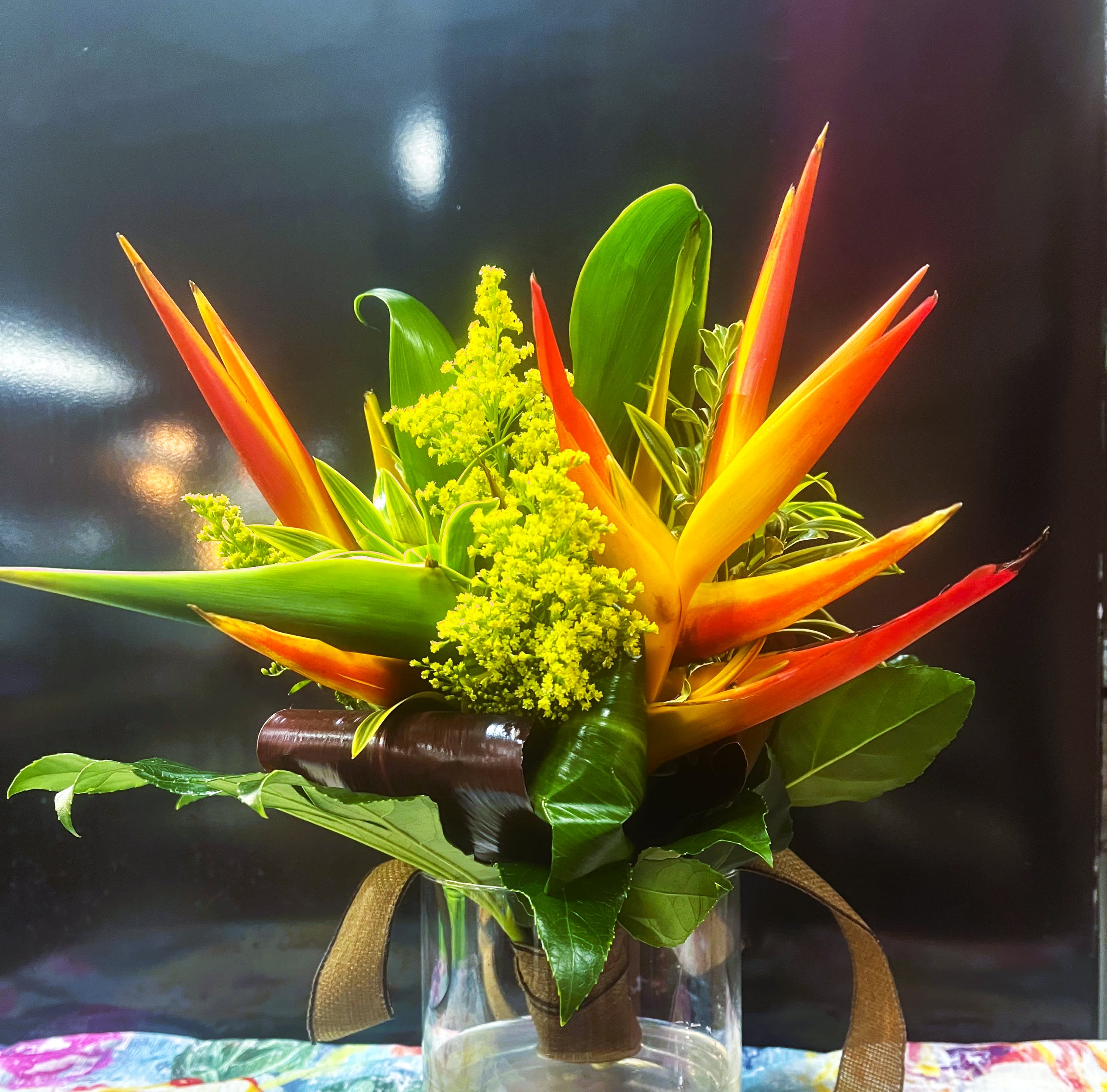 Compact tropical arrangement with orange and yellow flowers and bold green leaves in a clear glass vase.