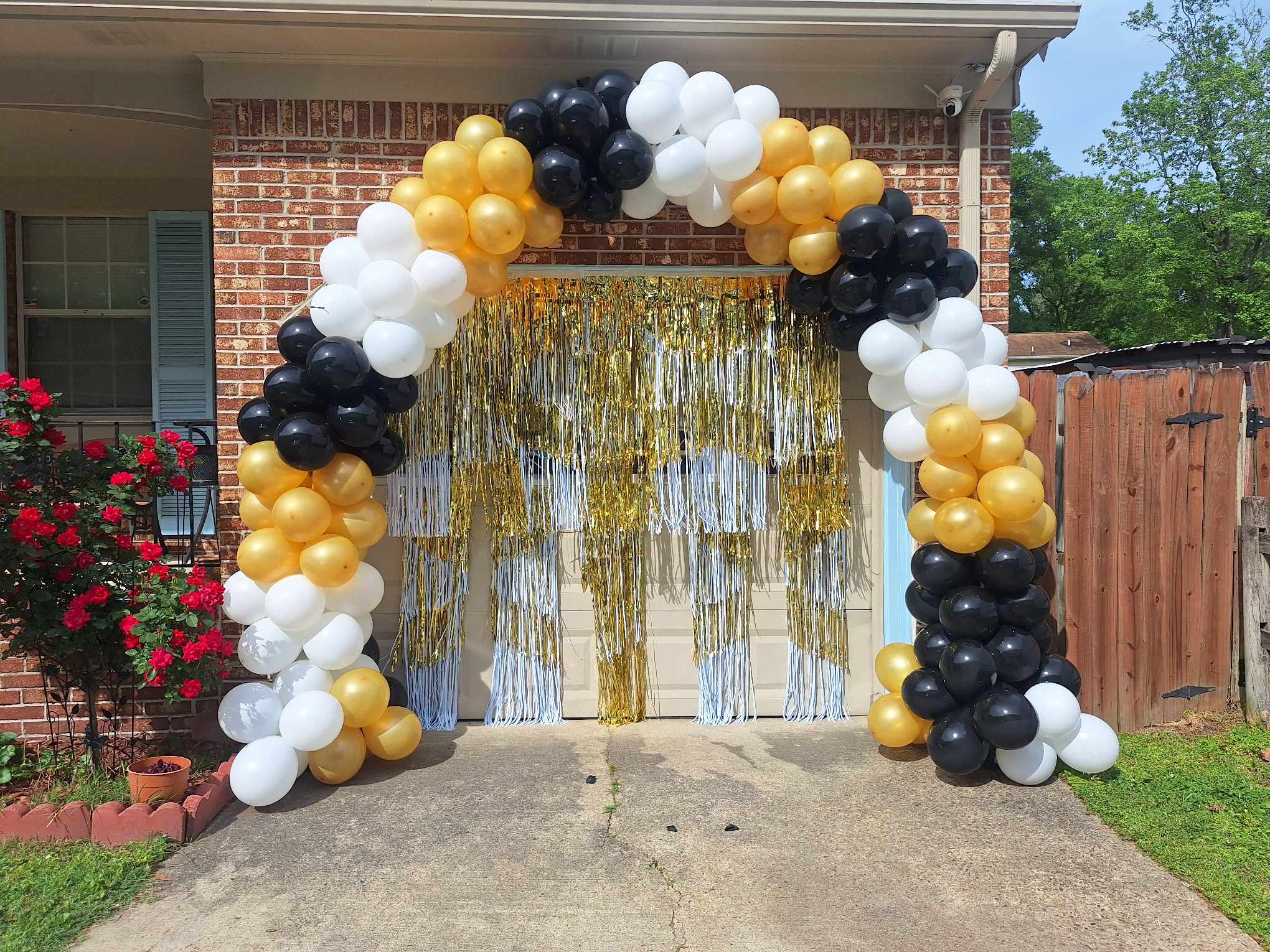 Black, white, and gold balloon arch framing a garage doorway with gold metallic fringe curtains.