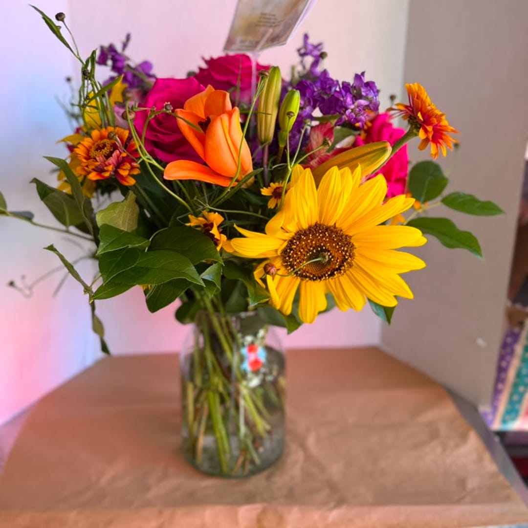 Mixed bouquet with sunflowers, roses, lilies, and purple blooms in a glass vase