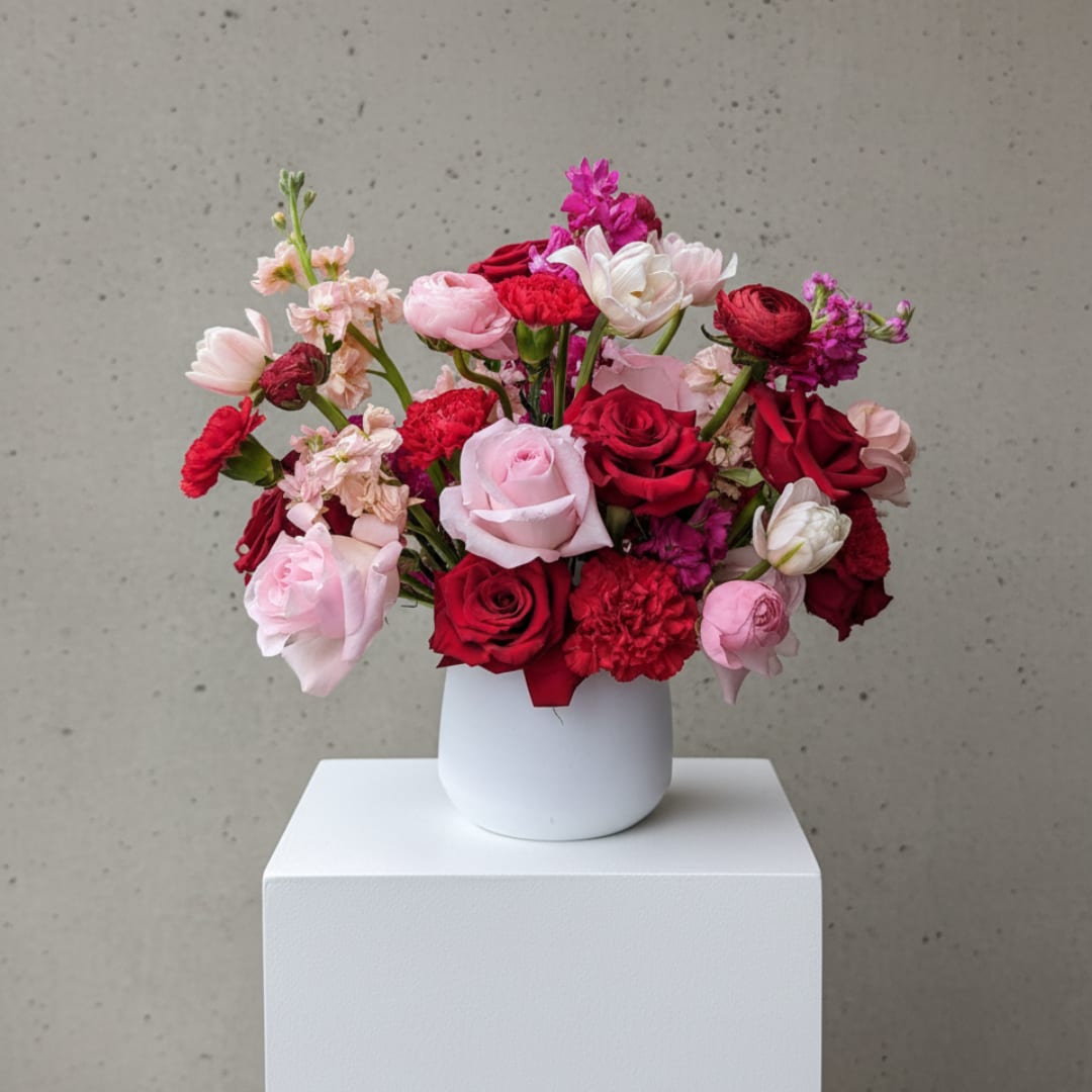 Velvet Kiss - Velvet kiss is a lush, garden-inspired arrangement filled with rich red, blush pink, soft ivory, and vibrant fuchsia blooms. Designed with roses, ranunculus, stock, carnations, and other premium seasonal flowers, this arrangement combines romantic color with natural movement for an effortlessly luxurious look. Presented in a modern white vessel, Velvet Romance is perfect for anniversaries, birthdays, romantic gestures, or simply making someone feel extra special.  Flower varieties may vary slightly based on seasonal availability while maintaining the overall color palette, style, and value shown.