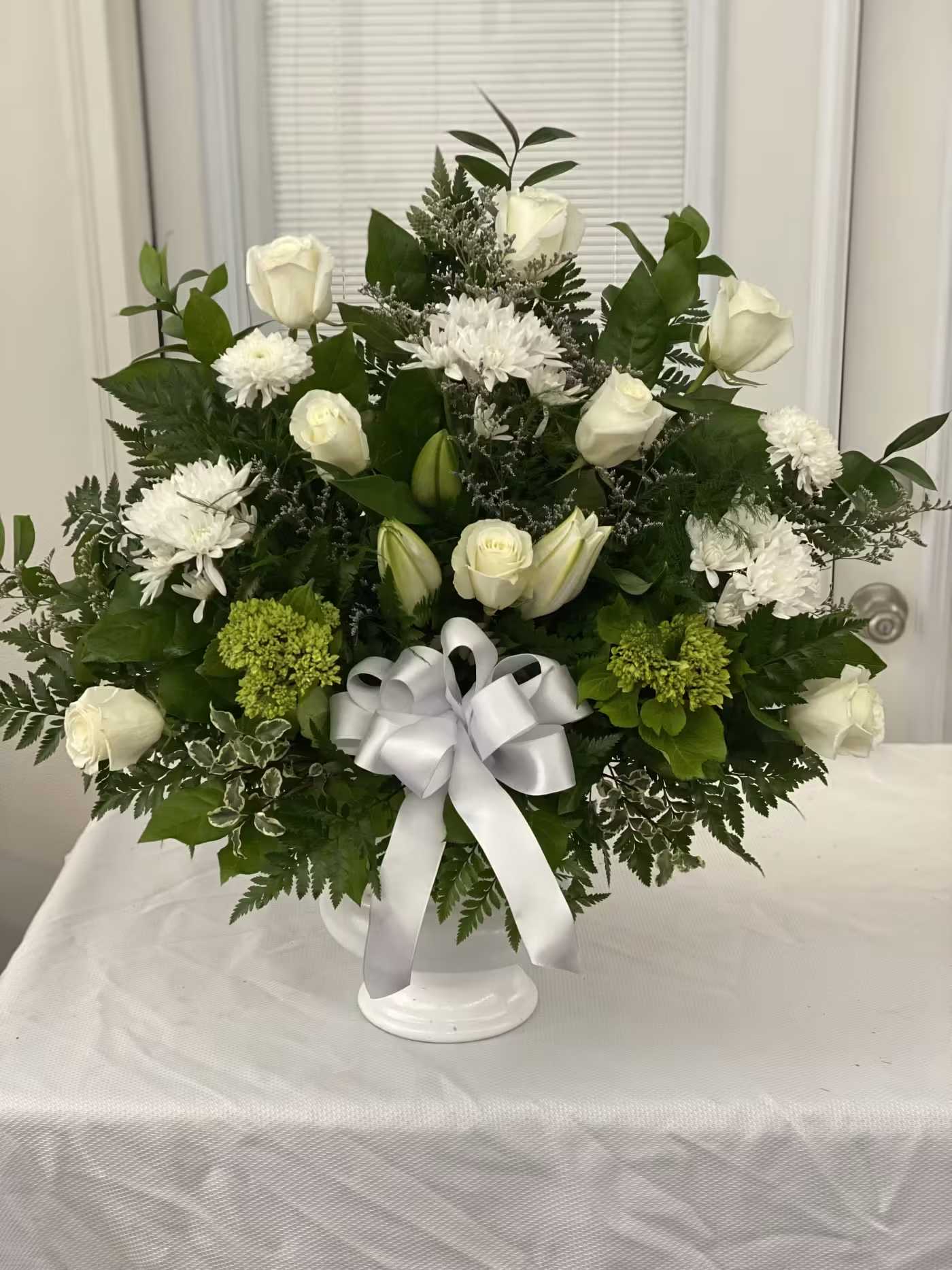 White roses and chrysanthemums arranged in a white vase with a silver bow