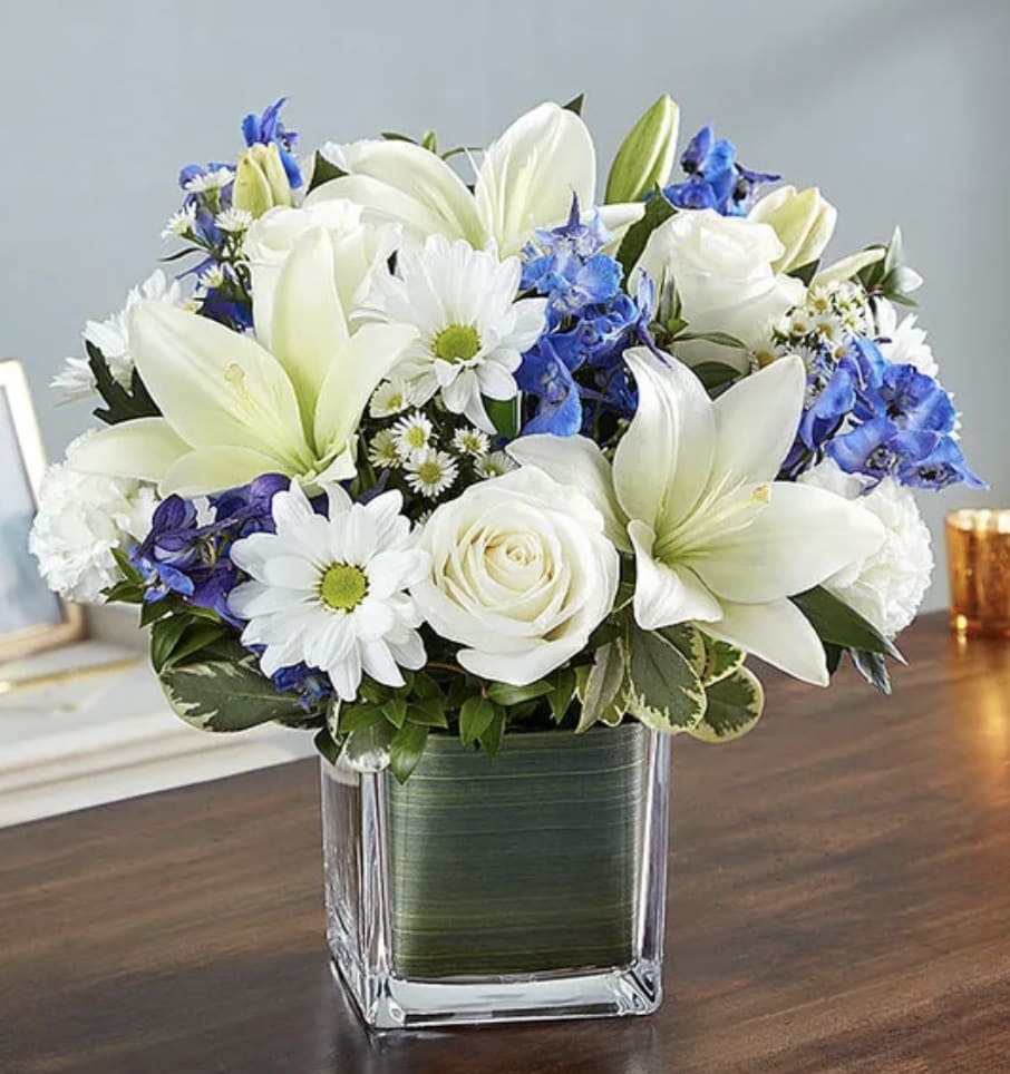 White lilies, roses, and blue flowers arranged in a square glass vase