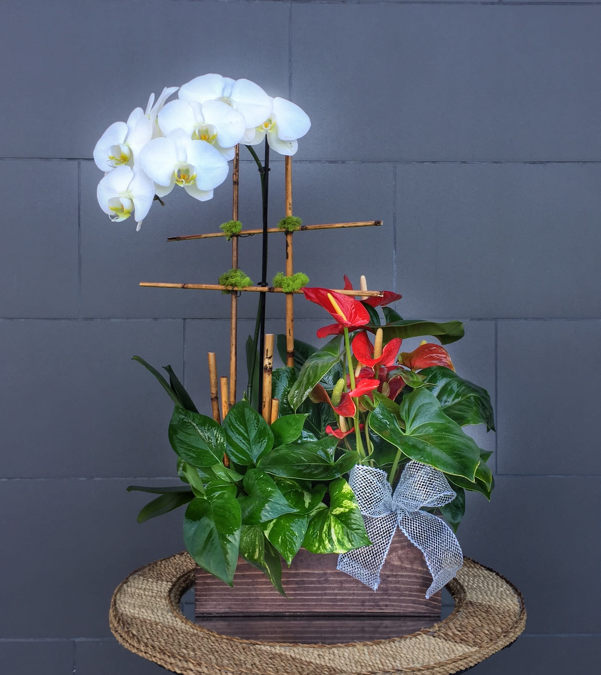 White orchids and red anthuriums in a wooden planter with a ribbon bow