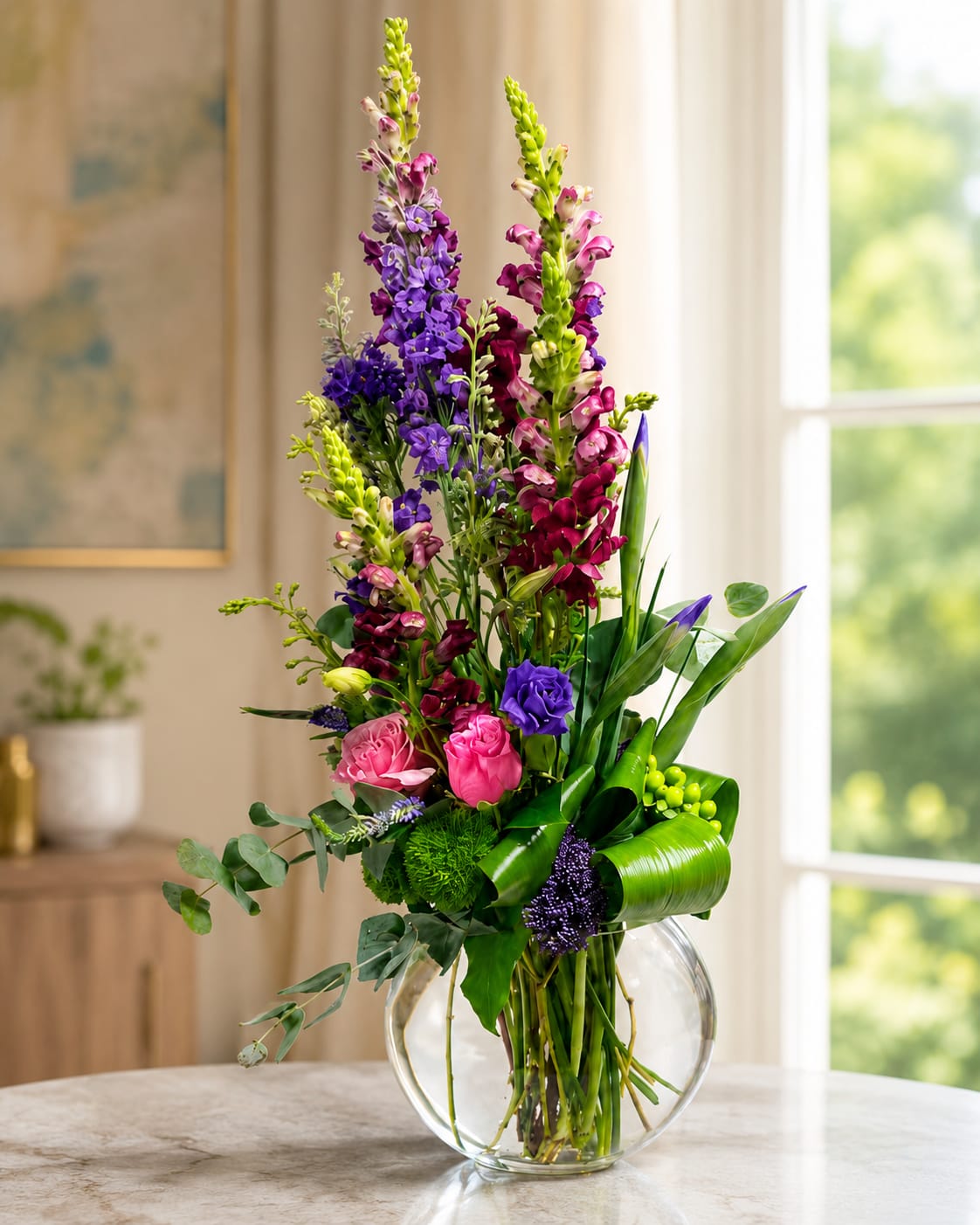 Chloe‘s Love by Petal Perfection  - Tall spires of purple and magenta snapdragons reach upward in this vibrant flower arrangement, creating an energetic, modern statement perfect for a birthday bouquet, Mother's Day gift, anniversary roses alternative, or a bright "thinking of you" surprise. Chloe's Love is built to make the person you're thinking of feel celebrated-its vertical silhouette and lively color mix also suit spring and Easter decor without feeling seasonal-only.  At mid-height, bright pink roses and deep purple lisianthus form a lush, rounded cluster, complemented by soft chrysanthemums and small green button-style blooms for pops of contrast and texture. Sculpted broad leaves add architectural movement while keeping the design fresh and contemporary. Stems are thoughtfully arranged in a wide, round clear glass vase that grounds the composition and shows off the natural lines and stems.  This arrangement works beautifully for celebrations (graduation, promotion, housewarming), comfort moments (get well, sympathy), and festive holidays from Mother's Day to Christmas. Care is simple: refresh the water every 2-3 days, trim stems at an angle if needed, and keep away from direct heat to prolong bloom life-an uplifting gift that will make her feel truly seen.