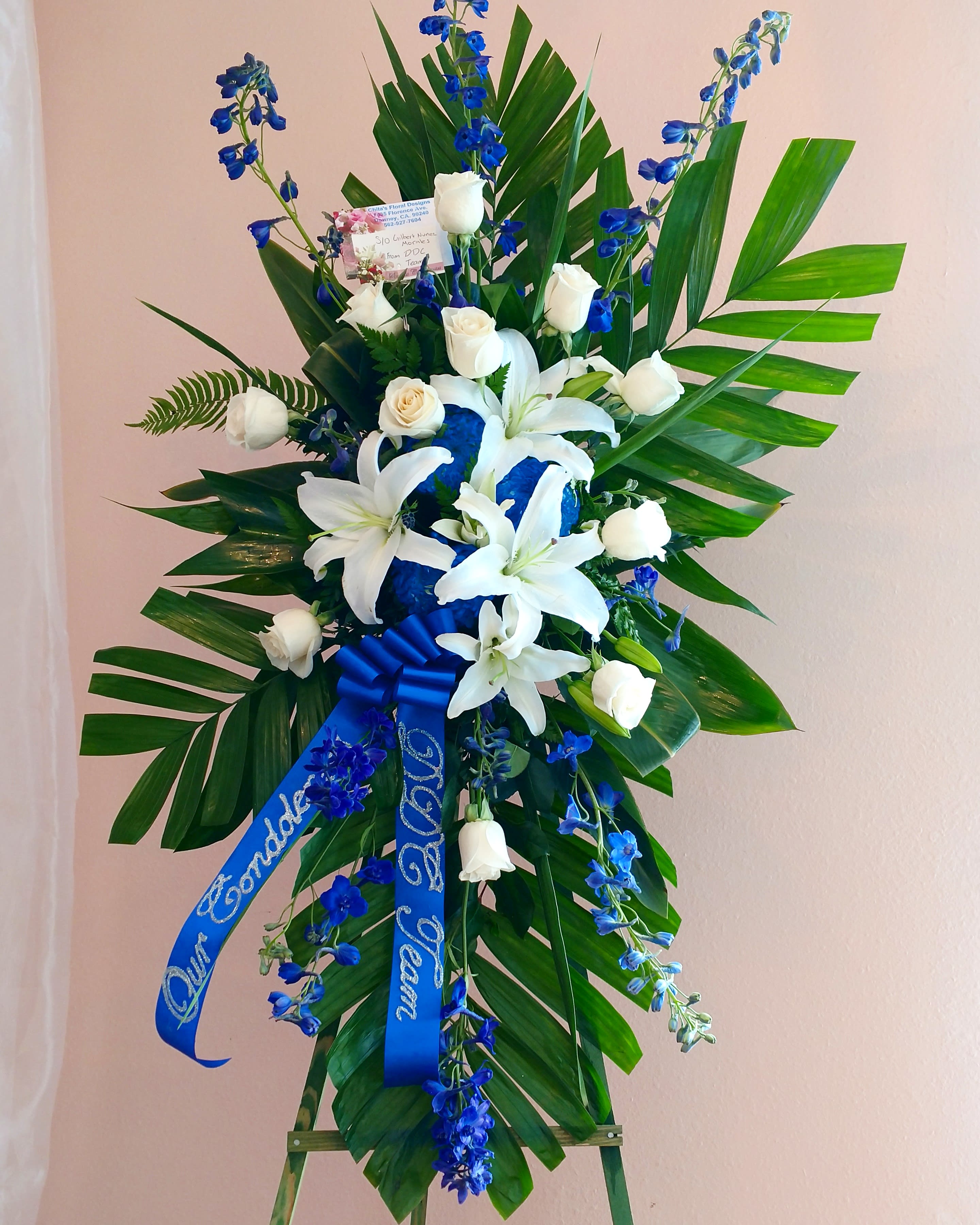 Tall standing floral spray with white lilies, white roses, and blue flowers on an easel.