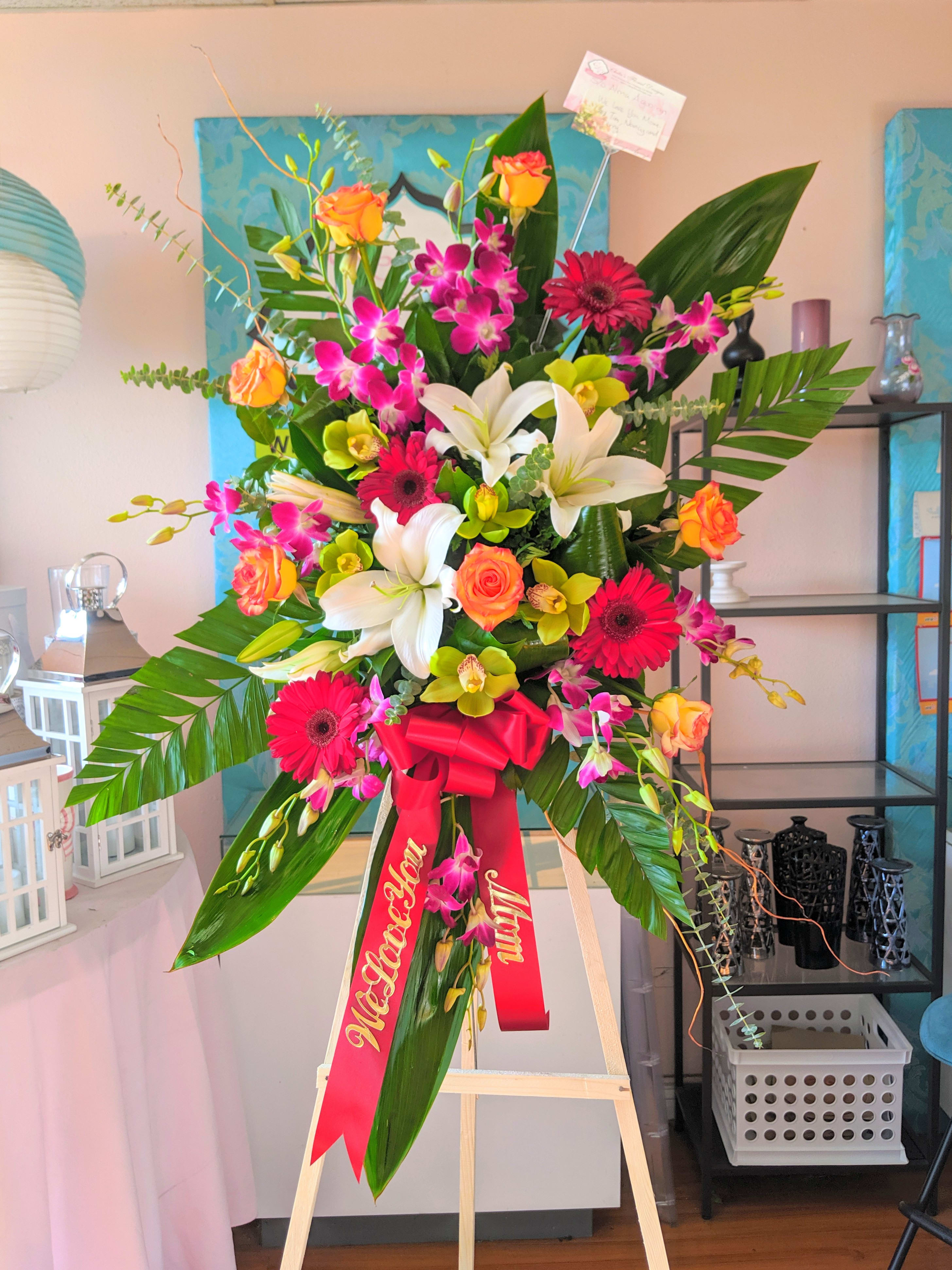 Tall standing floral spray with lilies, gerbera daisies, roses, and orchids on an easel