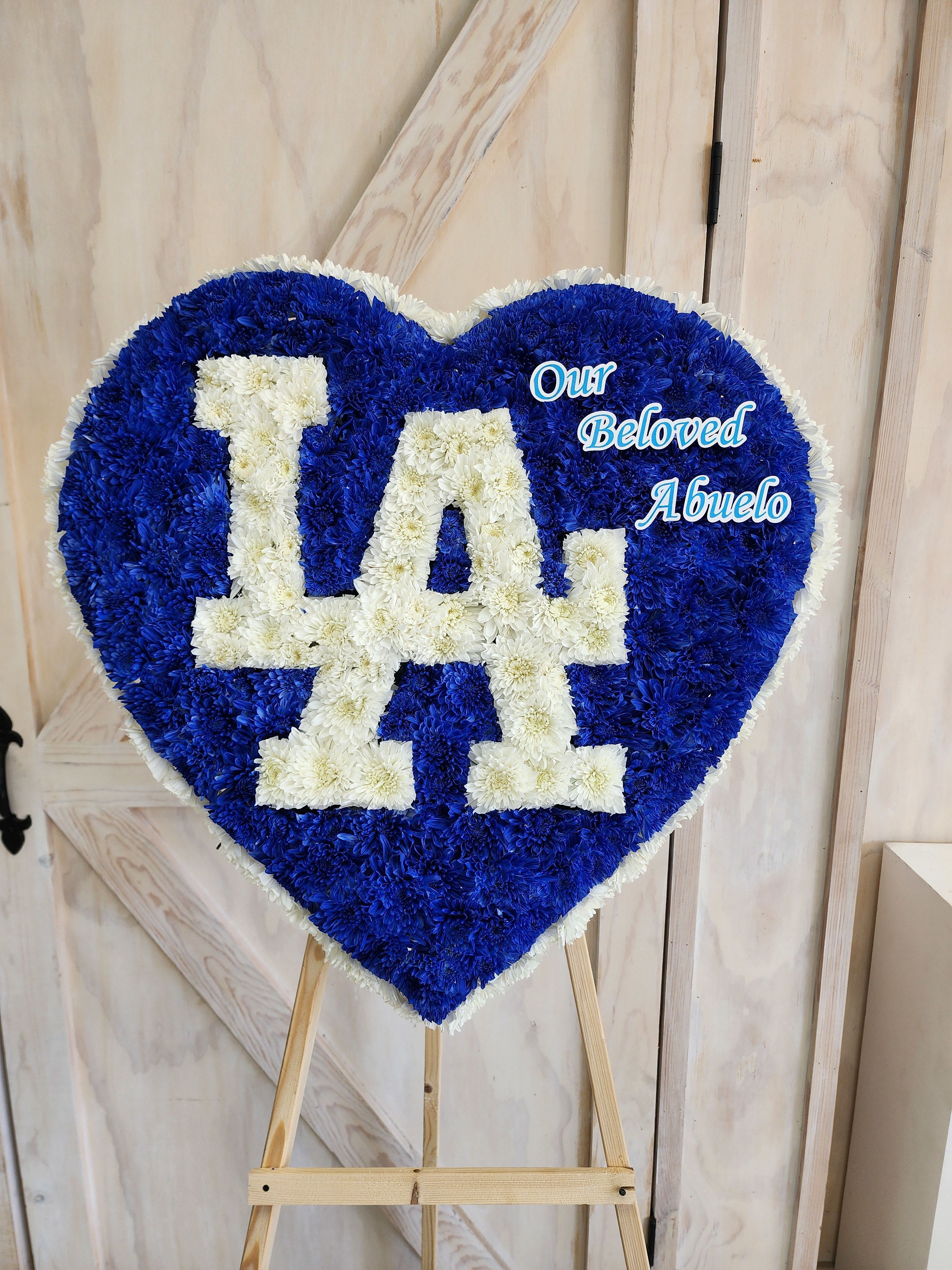 Heart-shaped floral tribute with blue and white flowers reading "LA" and "Our Beloved Abuelo"