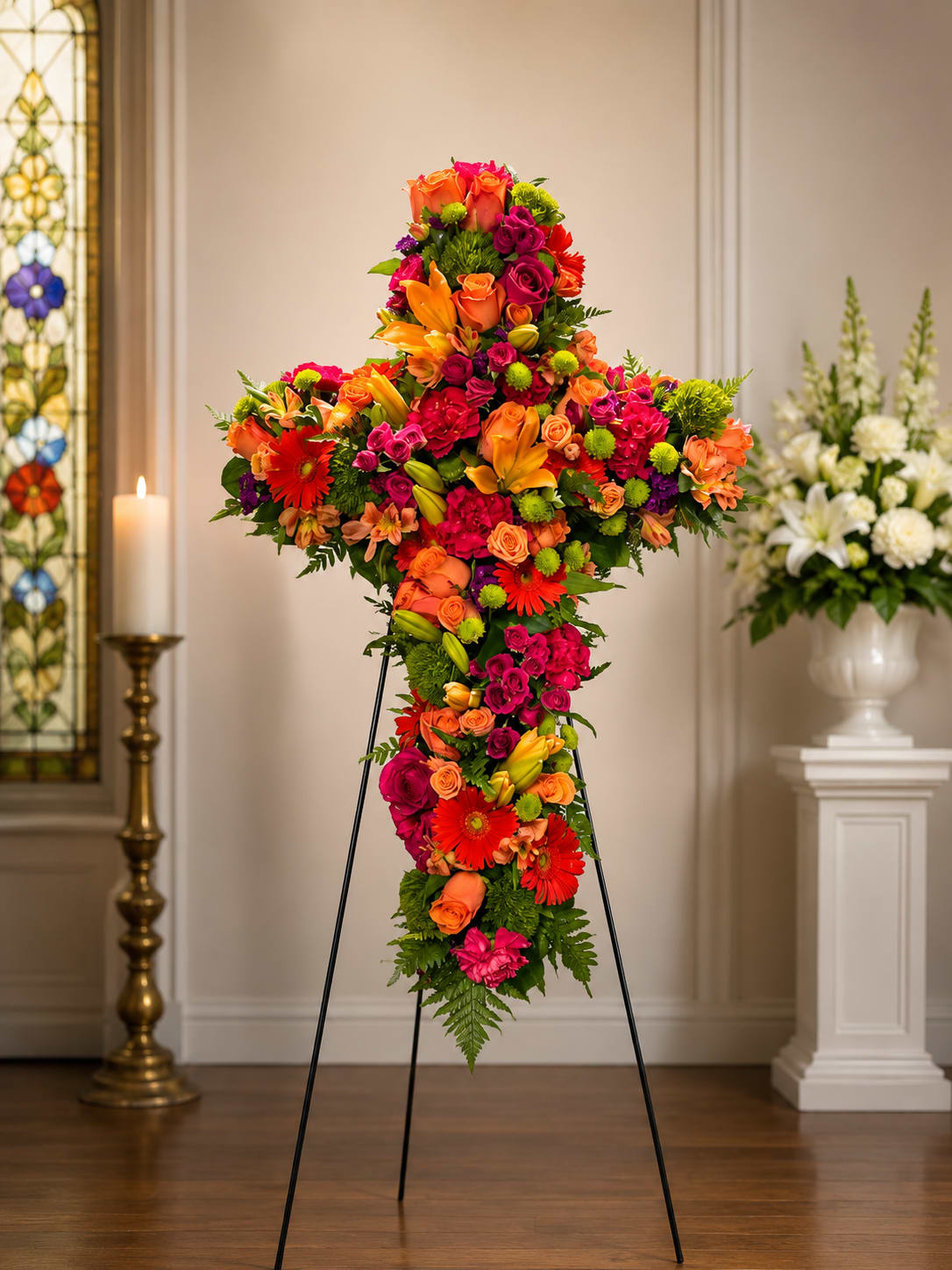 Citrus Enchantment Cross a Bryan Miller Jones Original  - A lush cross-shaped arrangement alive with orange, hot pink, red and lime-green accents-perfect as a sympathy or funeral tribute that feels uplifting rather than somber. This flower arrangement cross brings a bright, heartfelt presence to a service or church display and offers a comforting visual expression of faith and remembrance for the person you're honoring.  Carefully packed roses, Gerbera daisies, Asiatic lilies, spray roses and button chrysanthemums create a dense, textured surface with a tapered cascade that lengthens the silhouette for graceful impact. The palette includes touches of lavender, peach, yellow and subtle blue tones woven among the primary orange and pink, giving the piece depth and warmth. Sized to stand prominently at a memorial or to complement a sanctuary setting, the design arrives ready to display; keep flowers in a cool place away from direct sun and refresh water or floral foam as needed to extend bloom life.
