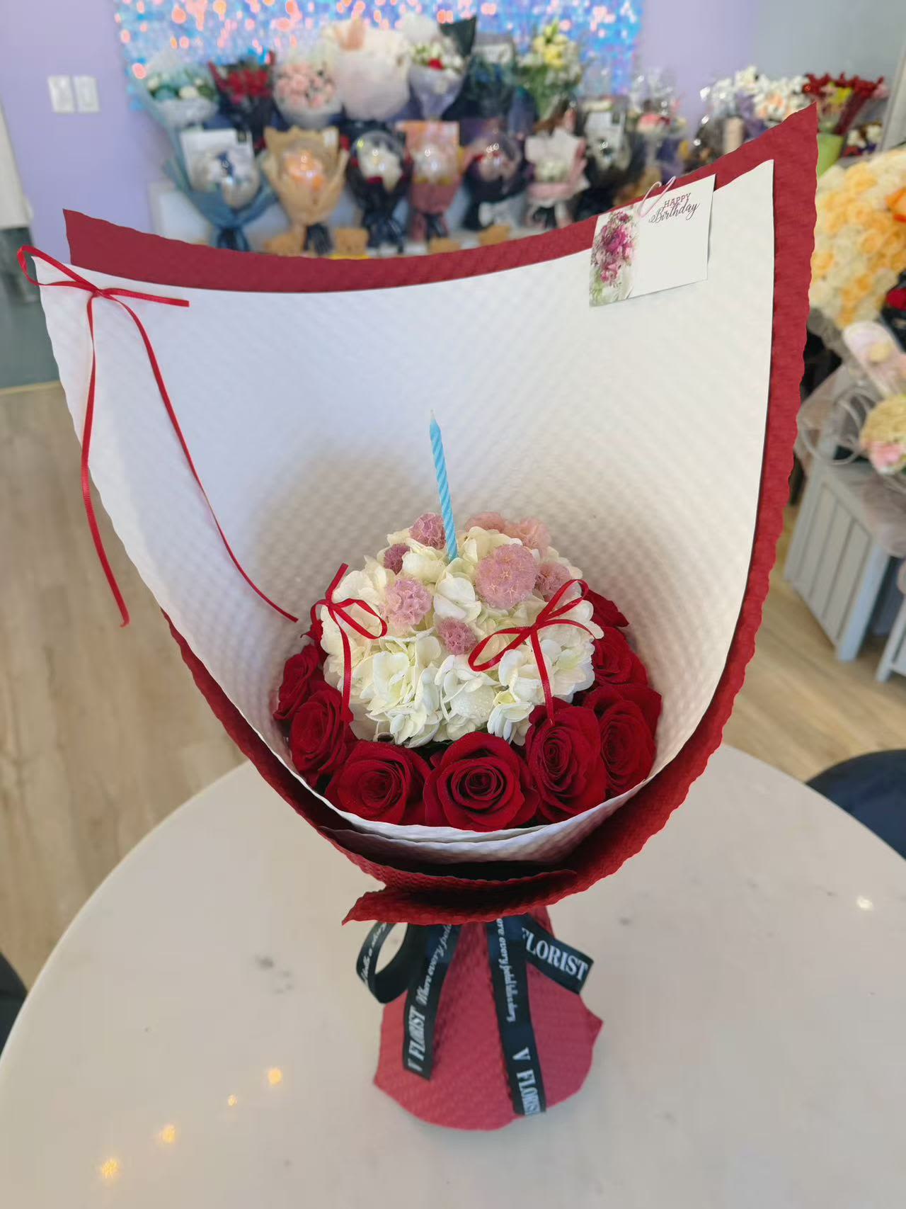 Birthday Bouquet - Celebrate a birthday most sweetly with this cake-inspired flower bouquet. This charming bouquet is designed to look like a mini birthday cake made entirely of fresh flowers. A ring of luxurious red roses surrounds a soft centerpiece of hydrangea. 