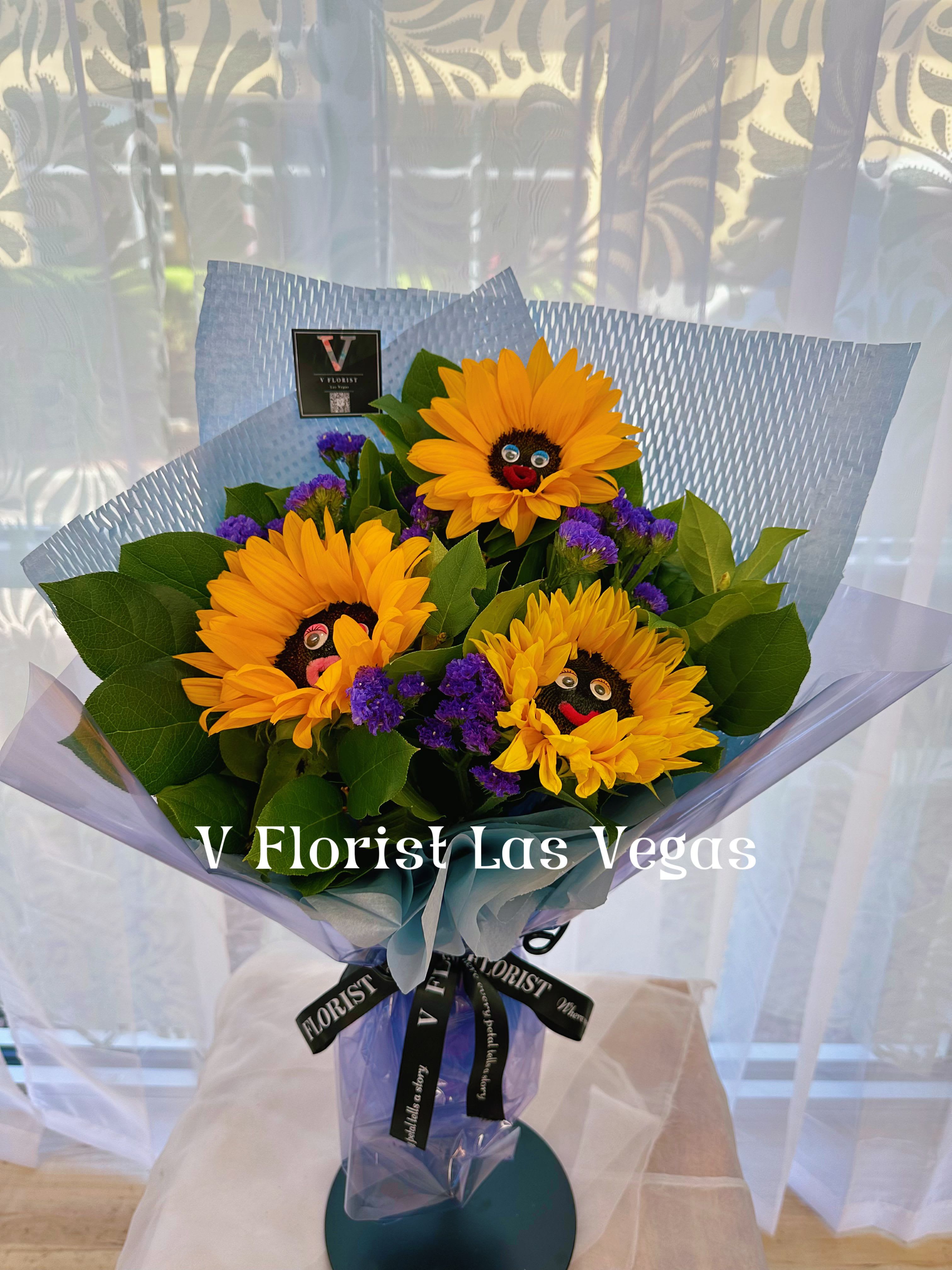 Hand-tied bouquet of yellow sunflowers with purple accents in blue wrap on a stand
