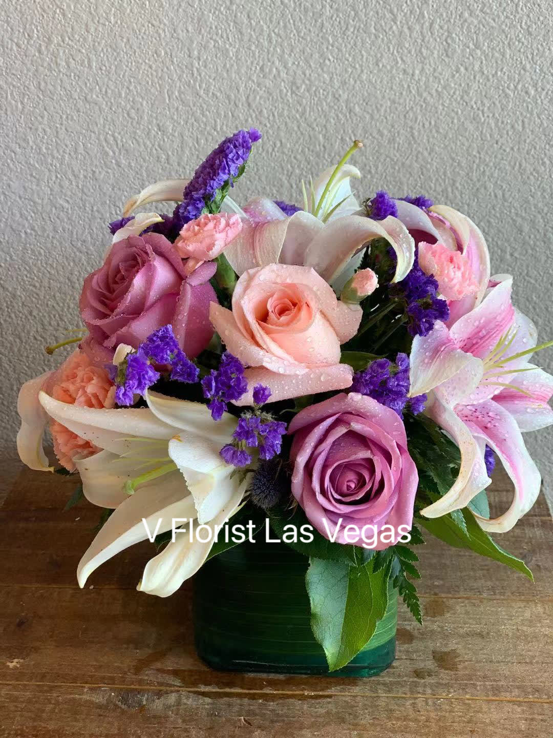 Compact arrangement of pink roses, lilies, and purple flowers in a green glass cube vase