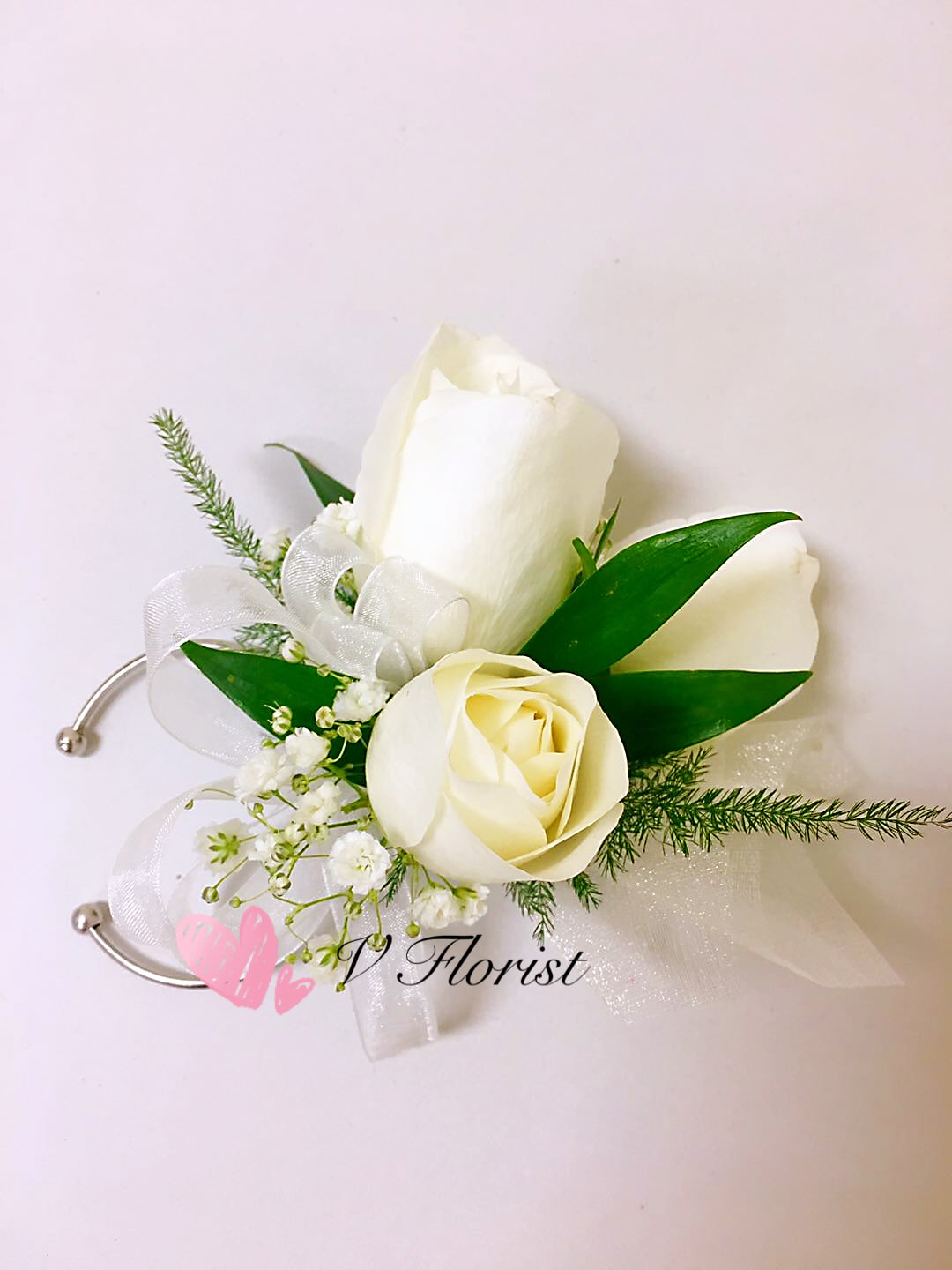White rose wrist corsage with small white filler flowers and sheer ribbon on a silver cuff.