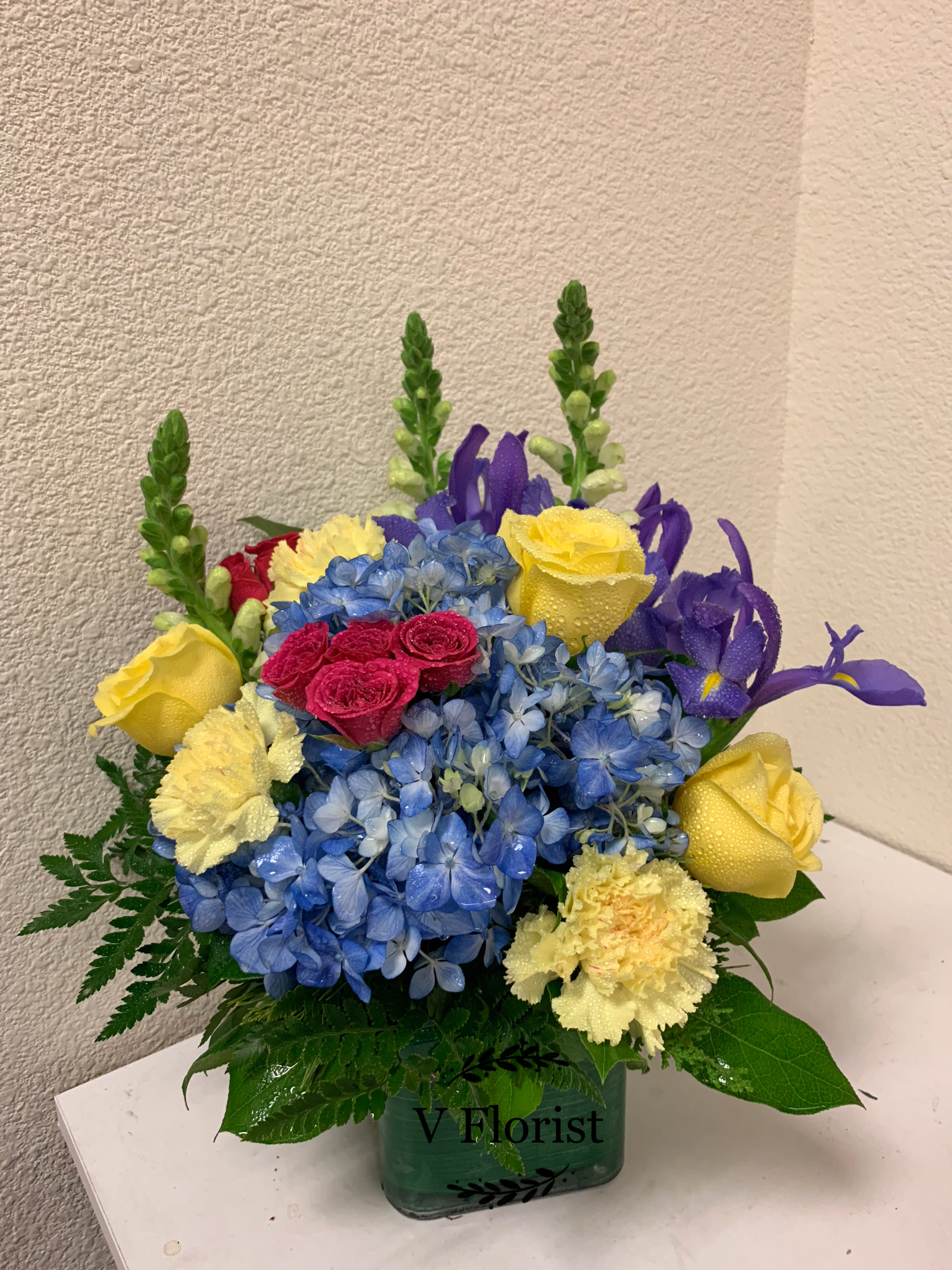 Bouquet of blue hydrangeas, yellow roses, and purple irises in a square vase