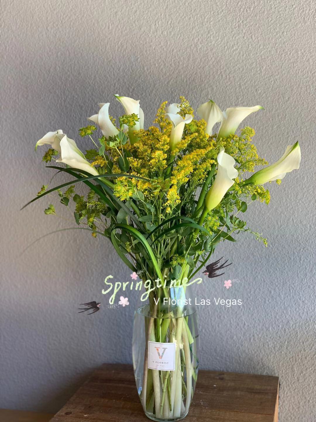 White calla lilies and yellow filler flowers in a clear glass vase