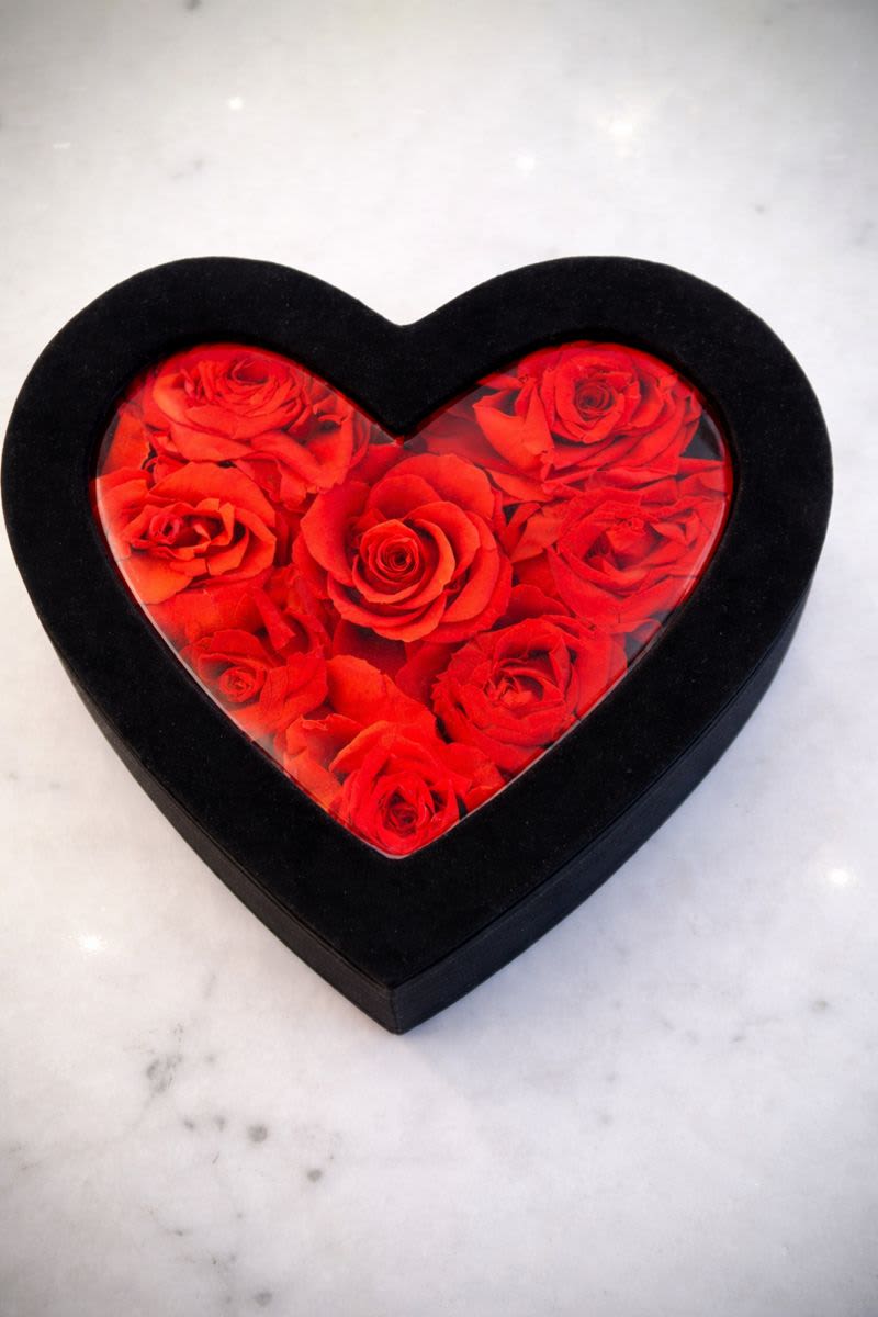 Heart-shaped black box filled with red roses under a clear lid on a light surface
