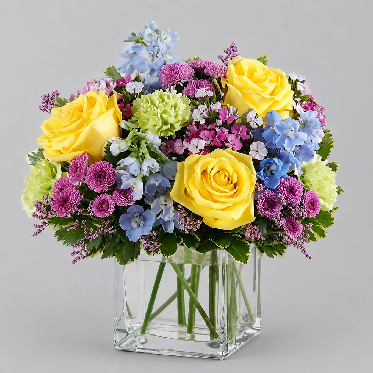 Mixed bouquet of yellow roses, blue flowers, and purple blooms in a clear glass vase