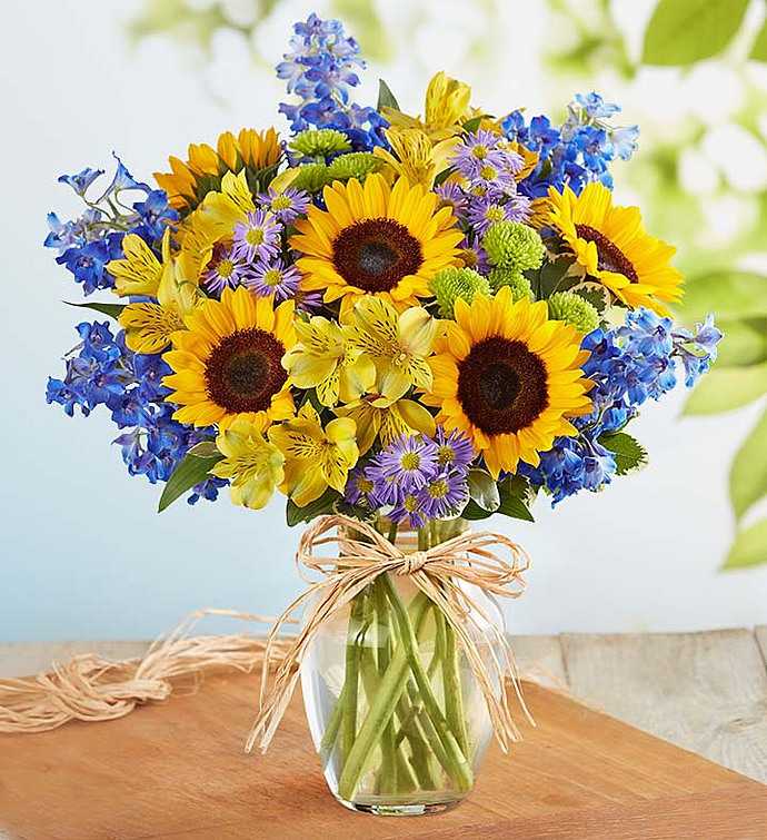 Spring Countryside - Inspired by the natural charm of the English countryside, our bright, seasonal arrangement of fresh sunflowers, delphinium, and more is hand-gathered inside a clear glass vase tied with raffia! It’s a must-have rustic design with European flair!