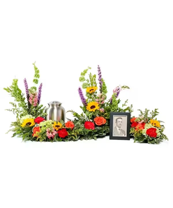 Joyful Sentiments of Color Urn Arrangement - Celebrate the joy your loved ones brought to your life while holding onto their memory with a colorful arrangement to envelop their urn and framed photographs. This display trails on a table to showcase the eternal love for those who have passed. Featured Blooms: Carnations, Roses, Snapdragons, Petite Sunflowers, Hydrangeas.   To guarantee the freshest bouquet possible, we may replace some stems in your arrangement for color or flower variety. While we always do the best to match the picture shown, sometimes different vases may be used. Any substitution made will be similar to the original design and be of equal or greater value.