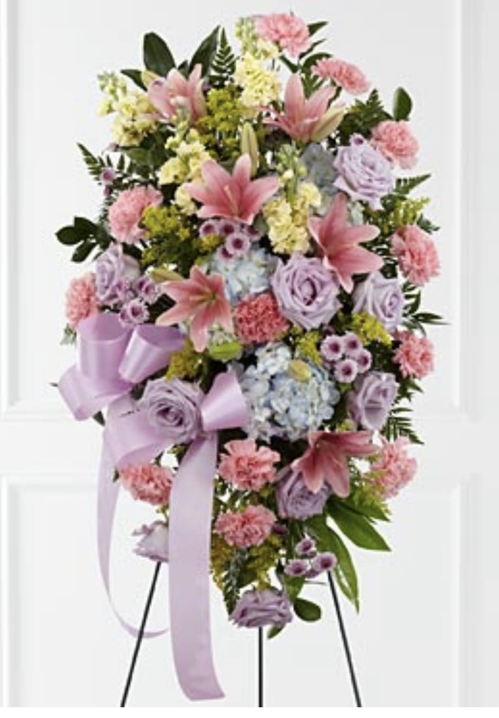 Standing spray of pink lilies, carnations, lavender roses, and blue hydrangeas with lavender ribbons on an easel.
