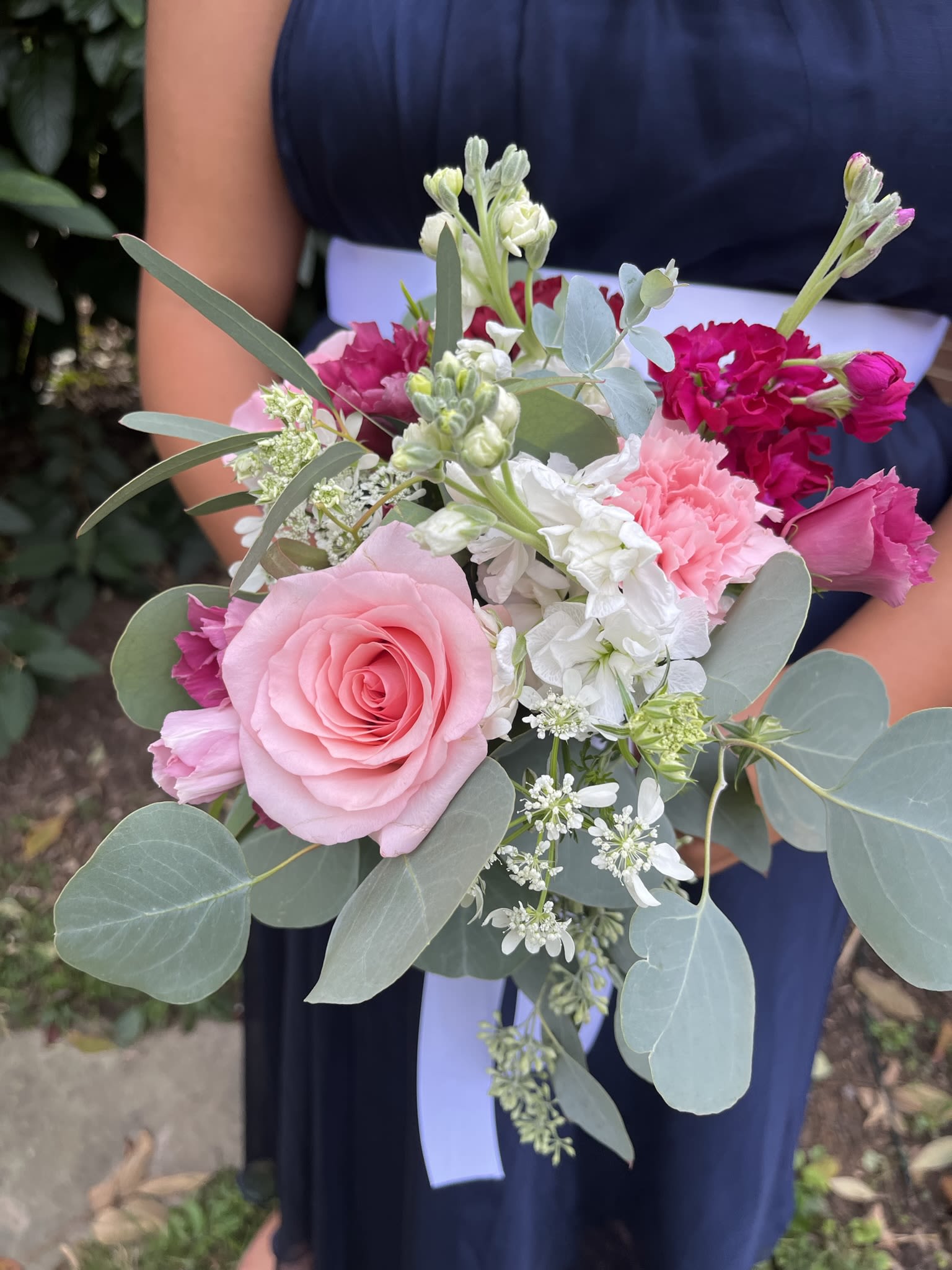 Hand-tied bouquet of pink roses, carnations, and white blooms with eucalyptus, held against a navy dress.