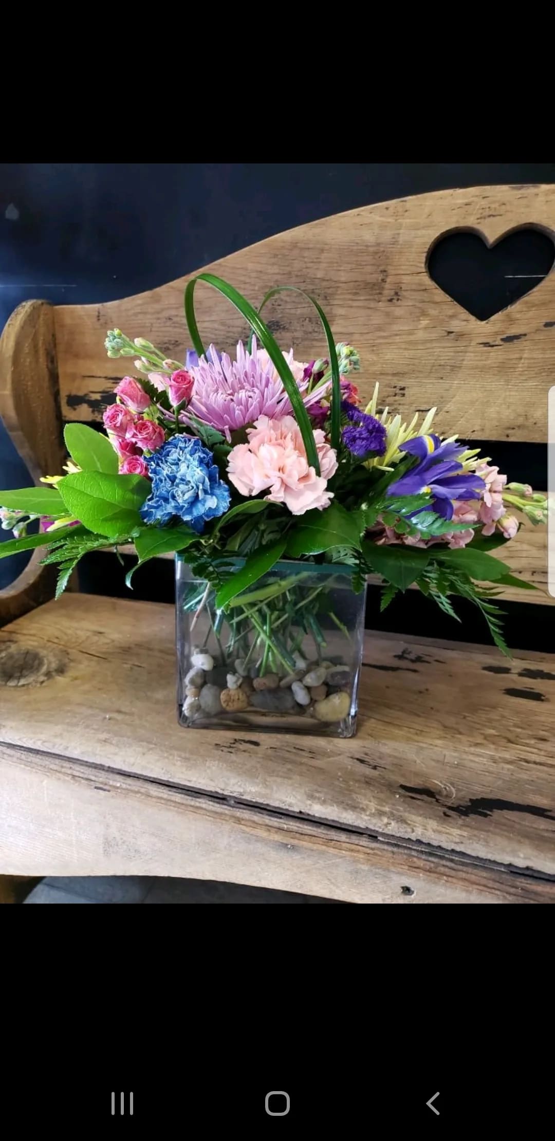 Rivers Rain - A low, elongated floral design that reads like a cheerful riverside bouquet-perfect as a birthday centerpiece or a "just because" surprise. This mixed-color flower arrangement brings together pink carnations and spray roses with lavender spider chrysanthemums, blue and purple blooms, and soft yellow accents to create a lively, uplifting display that works for get well wishes, new baby celebrations, or a romantic "thinking of you" moment.  Arranged in a clear glass cube vase and anchored with decorative river rocks, the compact profile makes it ideal for a bedside table, desk, or dining table. Arched loops of decorative grass rise above the cluster to add movement and height without overwhelming the low, horizontal silhouette. The combination of textures-from the ruffled carnations to the spidery chrysanthemum petals-gives the bouquet a fresh, spring-to-summer feeling while remaining appropriate year-round.  Choose this arrangement to say thank you, apologize with warmth, congratulate a friend, or brighten a boss's or admin professional's day-she'll feel genuinely seen when these bright, mixed blooms arrive. Care is simple: keep water fresh, trim stems if needed, and place out of direct heat to extend the life of the flowers.