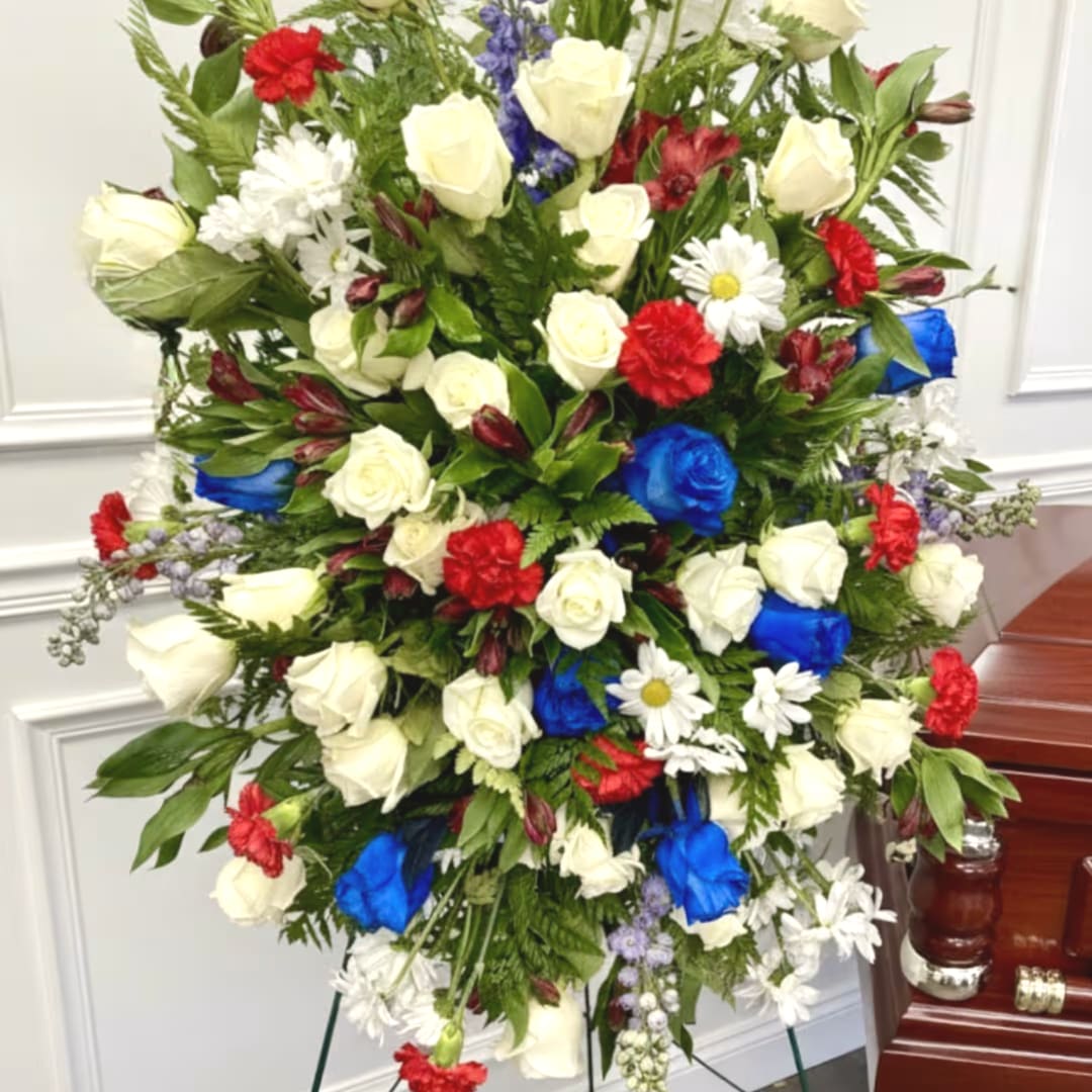 Stand &amp; Serve Easel   - A dignified standing spray that delivers a strong, respectful message-perfect as a memorial or funeral arrangement. This funeral standing spray blends white roses and white daisy-like blooms with vivid red carnations and blue-dyed roses to create a red, white, and blue palette that reads patriotic and ceremonial. Placed on a standing easel, its full, rounded silhouette gives immediate presence at a service so the person you're honoring feels remembered and celebrated.  The composition is dense and layered, built with abundant mixed greenery and a few trailing accents for texture and depth. Flowers included: white roses, red carnations, blue-dyed roses, white daisies, alstroemeria, and other seasonal blooms arranged on a sturdy standing easel. Its tall, upright form is designed to be seen from a distance and to complement traditional service settings.  Choose this arrangement when you want a classic, formal tribute that conveys respect and gratitude-especially fitting for veterans, patriotic ceremonies, or any service where a strong, composed display is desired. If you need delivery to a funeral home or help with personalization, contact us when ordering so we can confirm size and placement details.