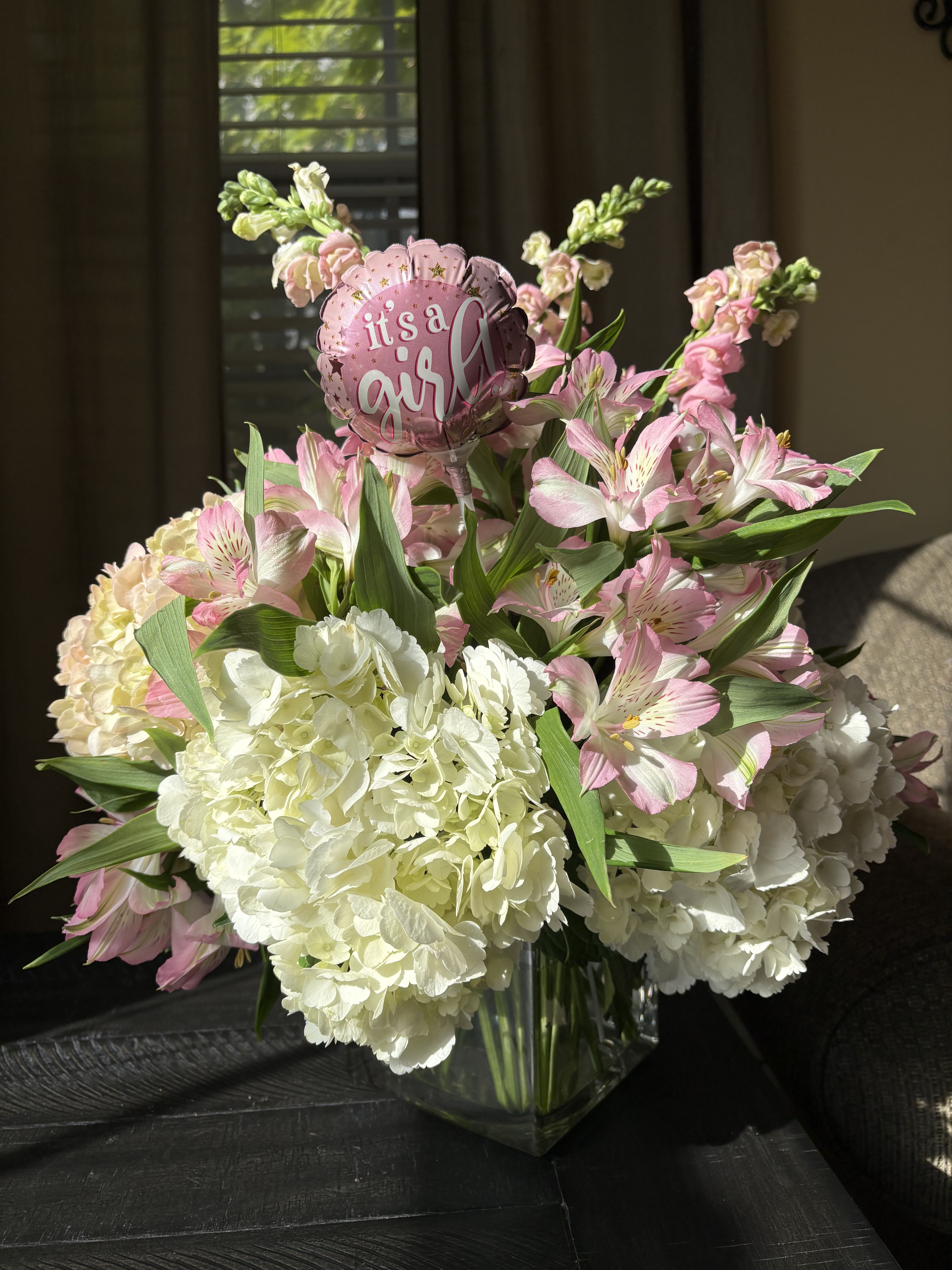 Pink and white flower arrangement in a glass vase with a "it's a girl" balloon