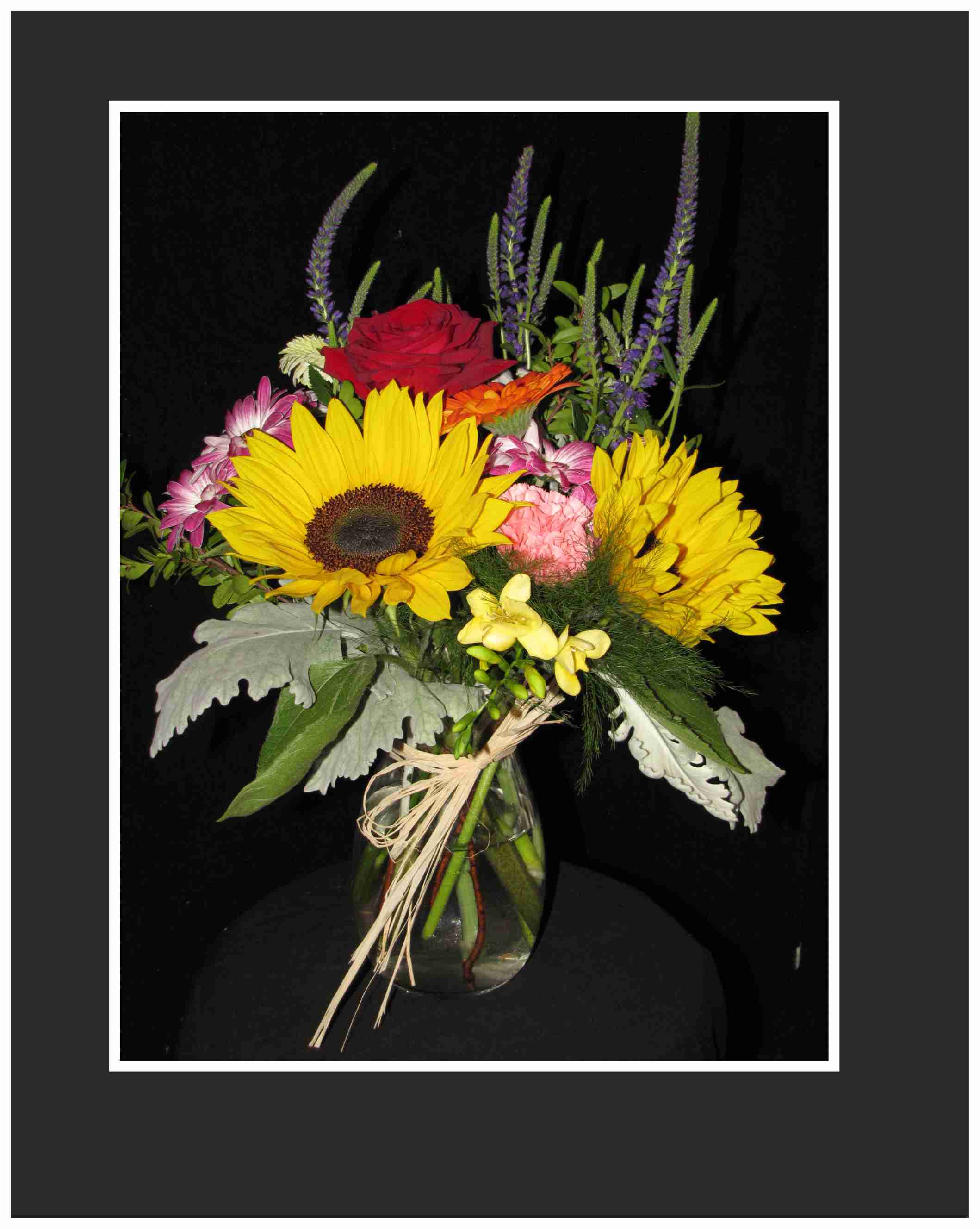 Happiness Begins Arrangement - An arrangement using all seasonal flowers sun flowers, gerbera daisies roses with freesia's and mixed greens arranged in an upright vase.  Approx.18 x 12 w