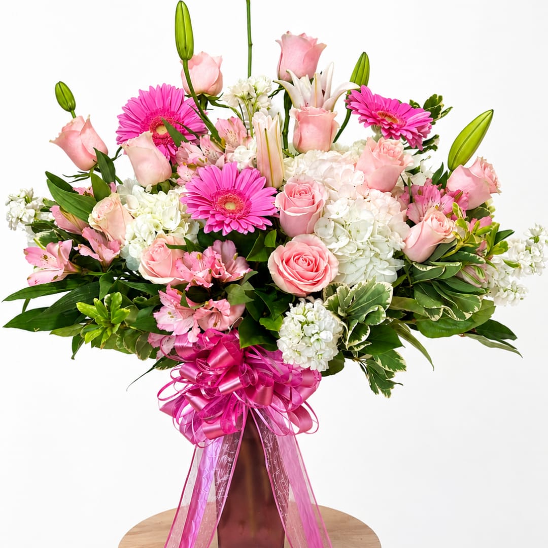 Pink roses, gerbera daisies, lilies, and white hydrangeas in a tall vase with a pink ribbon