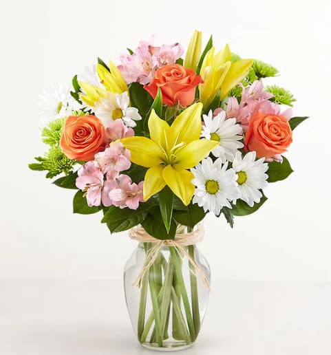 Bouquet of yellow lilies, orange roses, and white daisies in a glass vase