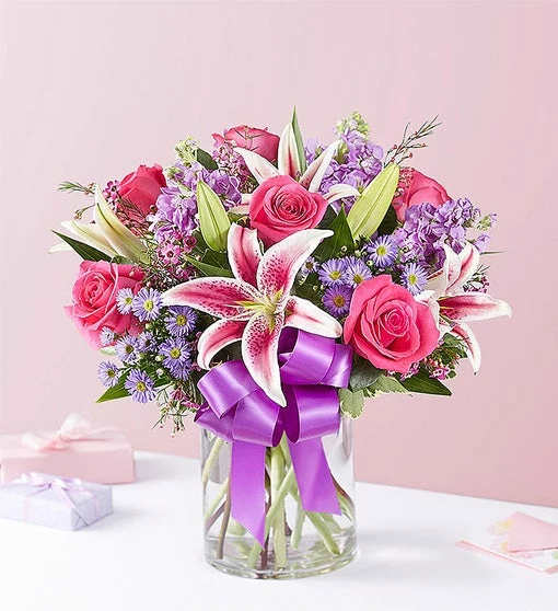 Pink roses and lilies in a glass vase with a purple ribbon