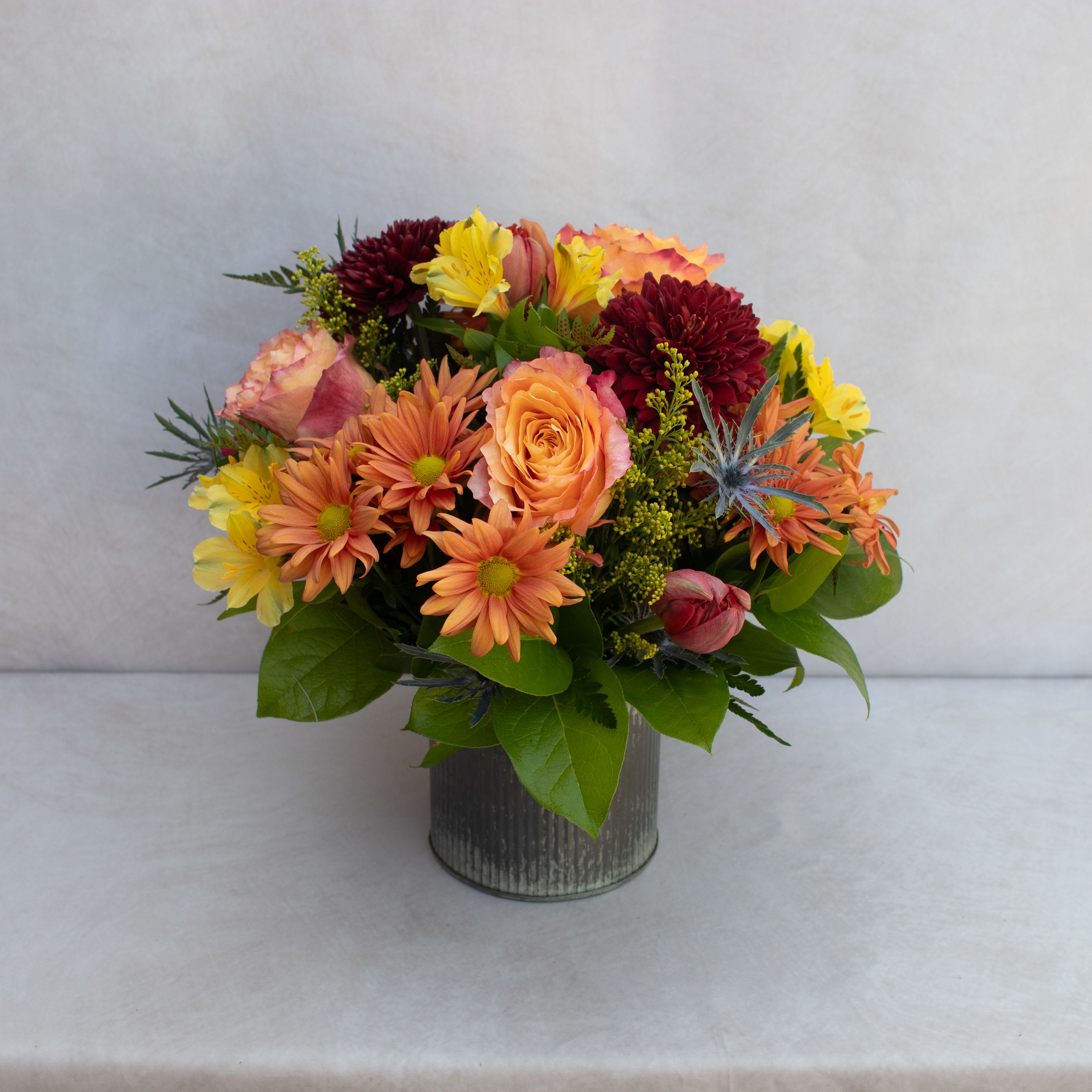 Mixed bouquet of orange, yellow, and burgundy flowers in a metal vase