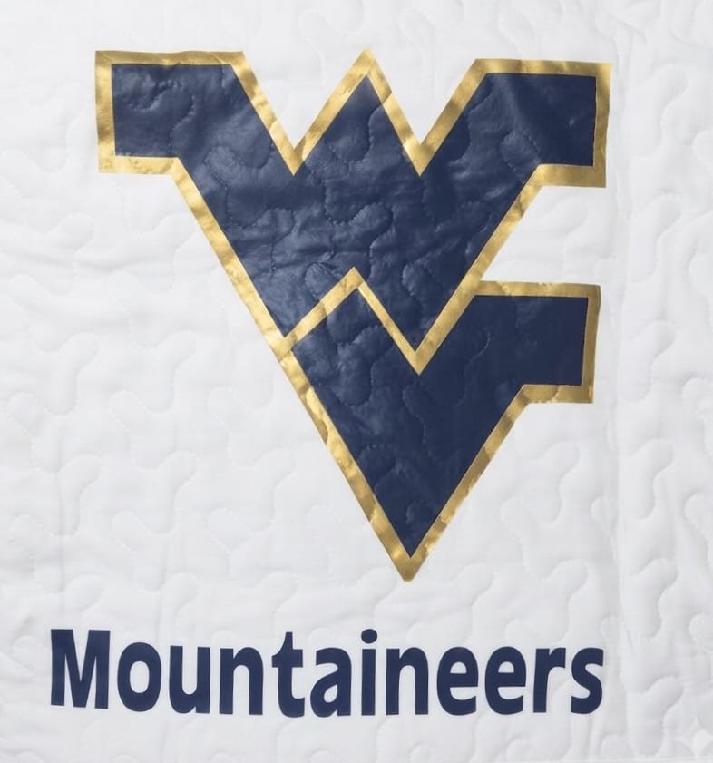 WV Mountaineers Quilted Throw  - A bold blue Mountaineers logo set against a crisp white quilted throw makes a spirited gift for the WVU fan in your life - perfect for game days, dorm rooms, or a thoughtful graduation present. This sporty, collegiate throw reads loud and proud and is ideal as a cozy keepsake to celebrate school pride or mark a special alumni moment.  Made from a white quilted textile with a large dark blue Mountaineers emblem outlined in metallic gold and the matching wordmark, the 50 x 60-inch throw brings strong graphic contrast and clean lines to any living space. It arrives flat and ready to drape over a couch, bed, or chair; pair it with the included wire easel stand for display at a tailgate, alumni event, or memorial tribute where a mounted presentation is desired.  Give the person you're thinking of a gift that shows you remember where their loyalties lie - warm, wearable team spirit that also looks great on display. The throw measures 50 x 60 inches.
