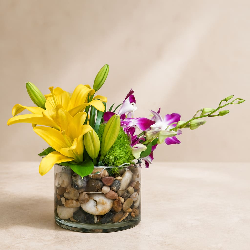 Yellow lilies and purple orchids in a pebble-filled glass vase