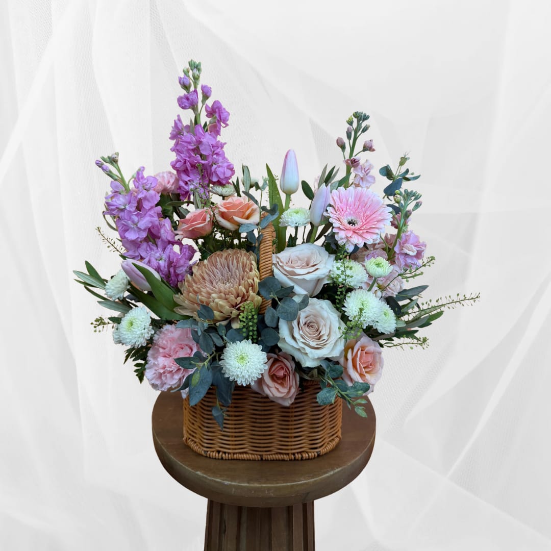 Pastel floral arrangement in a wicker basket with roses, gerbera daisies, and purple blooms