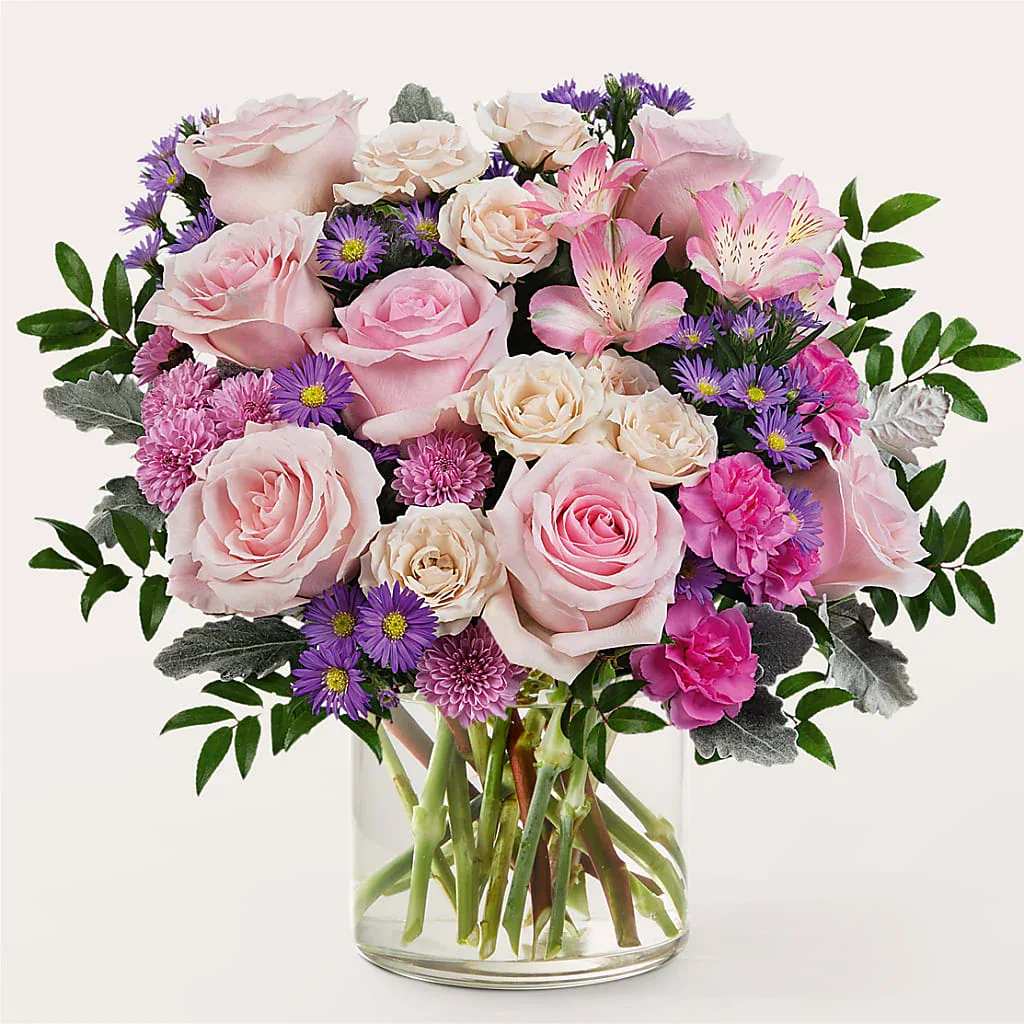 Always On My Mind - A lush cluster of blush and pink roses is the first thing the recipient will notice in this garden-style flower arrangement, making it a perfect birthday bouquet, anniversary surprise, or Mother's Day gift. The mix of purple daisy-like blooms and bright pink carnations adds cheerful contrast while pale cream flowers and alstroemeria keep the palette soft and romantic-ideal when you want someone to feel lovingly remembered.  Arranged in a clear glass vase that lets the stems show through, the design reads airy yet abundant, with layered blooms creating a full, rounded silhouette that looks polished on a dining table or desk. Colors: pink, purple, white. Flowers: roses, daisies, carnations, alstroemeria. The result is the kind of thoughtful bouquet that makes the person you're thinking of feel truly seen and celebrated-easy to order for birthdays, anniversaries, thank-you gestures, or just because.