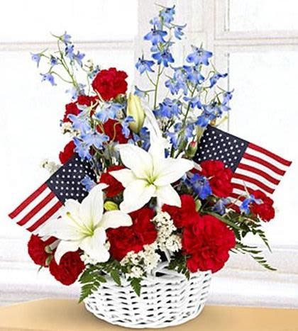 Blue and White Blossoms - Bright, ceremonial and unmistakably patriotic, this blue and white blossoms arrangement is a tasteful choice for Memorial Day, Veterans Day, or any occasion when you want to honor a loved one. The upright bouquet in a white wicker basket pairs red carnations, white lilies and tall blue blooms to create a bold red‑white‑blue statement that reads both respectful and celebratory - the kind of arrangement that makes the recipient feel honored and remembered.  The composition is full and structured, with the taller blue flowers rising above a dense cluster of red carnations and open white lilies for contrast and texture. Two small American flags are tucked into the design for a classic memorial or patriotic touch; the white wicker basket provides a clean, traditional base that suits indoor displays or graveside tributes. This arrangement arrives ready to display and conveys a clear, heartfelt message of remembrance and gratitude.