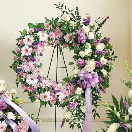 Lavender Tribute Wreath  - Sympathy wreath that reads as a gentle, heartfelt tribute-this Lavender Tribute Wreath is made for memorial services and occasions of remembrance. The floral arrangement combines pink, white and purple blooms with lush green foliage to create a soft, romantic look that comforts the person you're honoring and those who gather to remember them.  Clusters of daisies, roses and complementary seasonal blossoms are arranged densely around the heart form, with a lavender ribbon tied to a metal easel for dignified display. The pastel palette and full greenery deliver an elegant, understated presence that photographs beautifully and carries across the service space.  Perfect for funerals, memorials, or to leave at a graveside, this wreath is sized to stand on the included black metal easel and arrives ready to display. Light misting of the flowers and placement out of direct sun will help preserve their freshness during services.