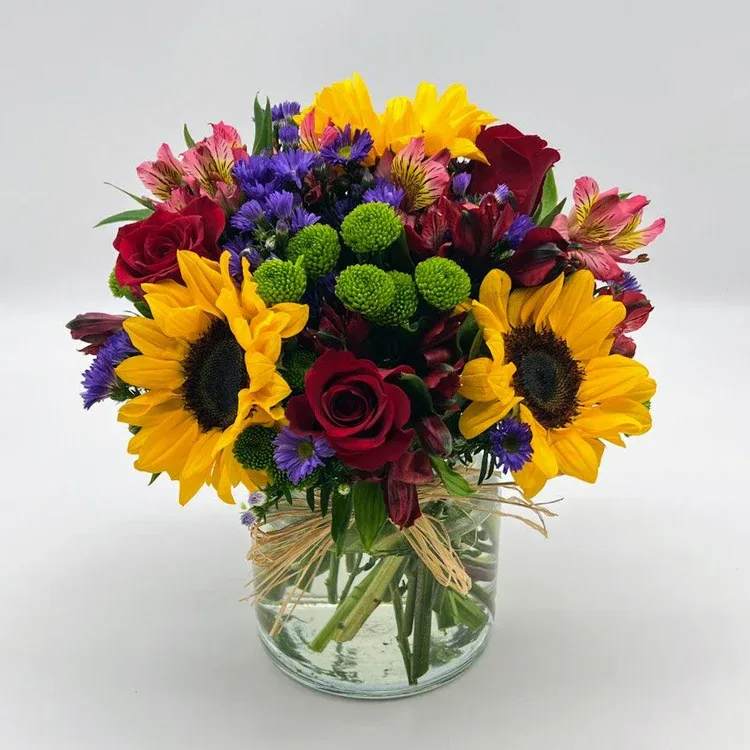 Beautiful Blooms  - Soft-white lilies and blushing pink roses mingle with clusters of purple stock and airy aster to create a romantic flower arrangement perfect for a birthday bouquet, anniversary roses alternative, or a thoughtful "just because" gift. The clear glass vase and large pink ribbon give the presentation a polished, feminine feel so the person you're thinking of will arrive at the moment feeling noticed and cherished.  Two style options appear from the images: one leans pastel and refined-white lilies, pink roses, purple filler, eucalyptus-like foliage, wrapped in pale paper and finished with a pink ribbon around a clear glass vase-while the other is bright and joyful with large yellow sunflowers, deep red roses, pink alstroemeria, purple filler, and green button chrysanthemums tied with raffia for a relaxed, hand-tied look. Both are full, rounded arrangements featuring chrysanthemums and other seasonal blooms for texture and contrast.  Either choice makes a confident statement for birthdays, anniversaries, thank-you gifts, or everyday celebrations; select the softer palette to convey gentle romance or the sunflower mix to spark cheer. The bouquet arrives arranged in a clear glass vase-no extra steps required beyond fresh water-so the recipient can enjoy the vibrant colors and layered textures right away.