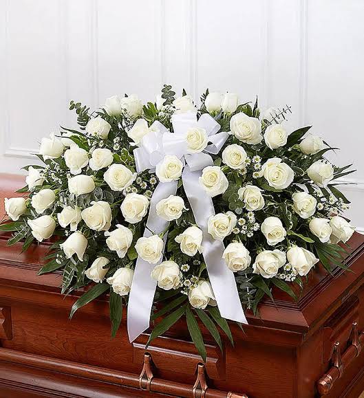Eternal Grace White Rose Casket Spray - A low, rounded casket spray of white roses that reads with quiet dignity-perfect as a sympathy or funeral tribute for someone you want remembered with grace. Placed directly on a wooden casket, the arrangement's dense clusters of roses and airy green fillers create a soft, restrained silhouette that comforts at a difficult moment.  Crafted to lie flat across the lid, this spray combines large white roses with pale pink rose accents and mixed greenery, accented by a wide white ribbon for a formal, ceremonial touch. The palette of white, pink, and green keeps the presentation tender and respectful while the spread covers the casket top for a balanced, calming effect.  Designed to express sympathy and honor-whether for a close family member or a cherished friend-this arrangement says "you are thought of" in a timeless way. Sized to rest gently on a standard wooden casket; minimal care is needed once placed, and our team prepares and delivers it ready for service.