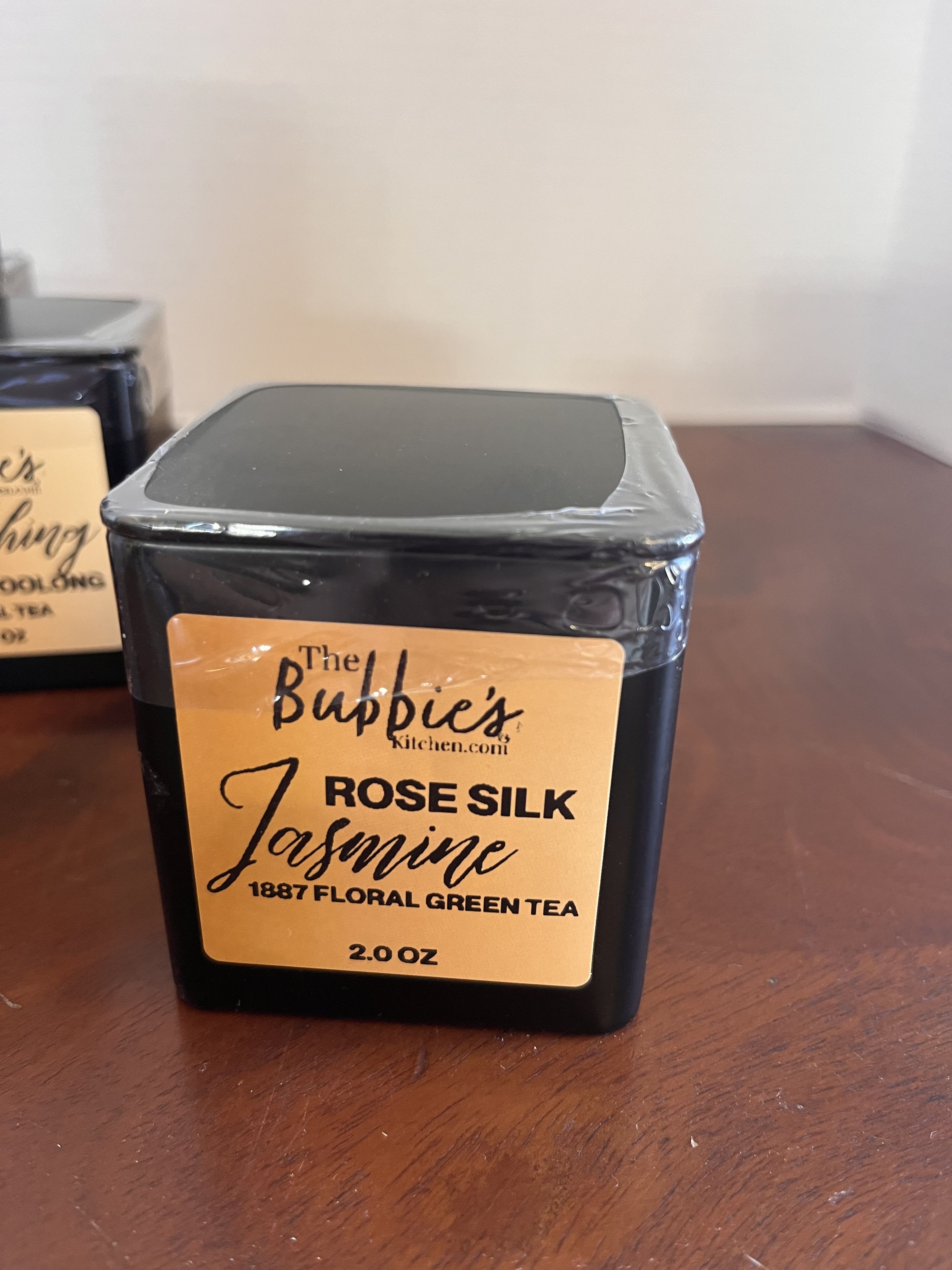 Square black tin of Rose Silk Jasmine floral green tea with gold label on a wooden surface