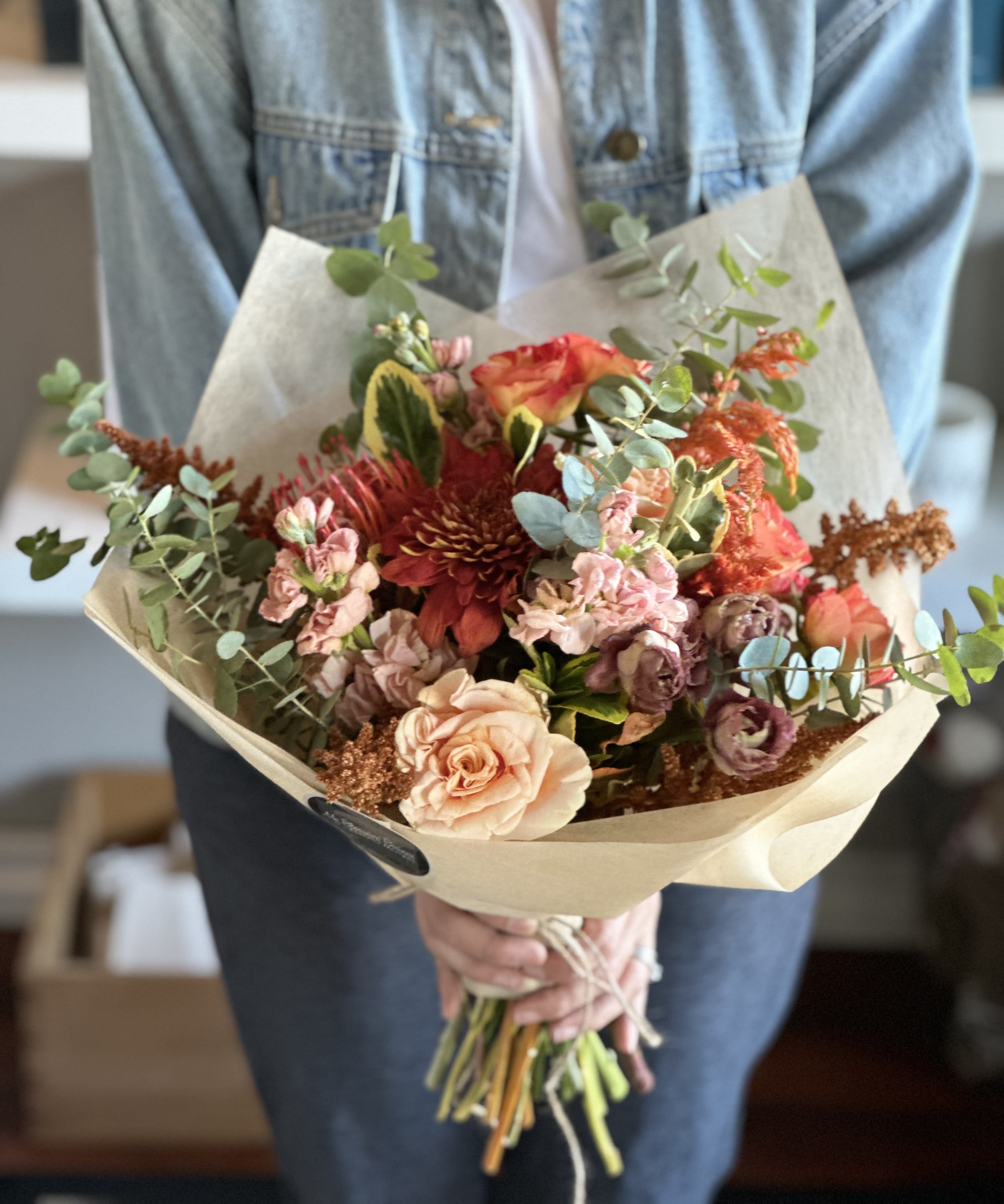 Handheld bouquet of peach, red, and pink flowers wrapped in paper