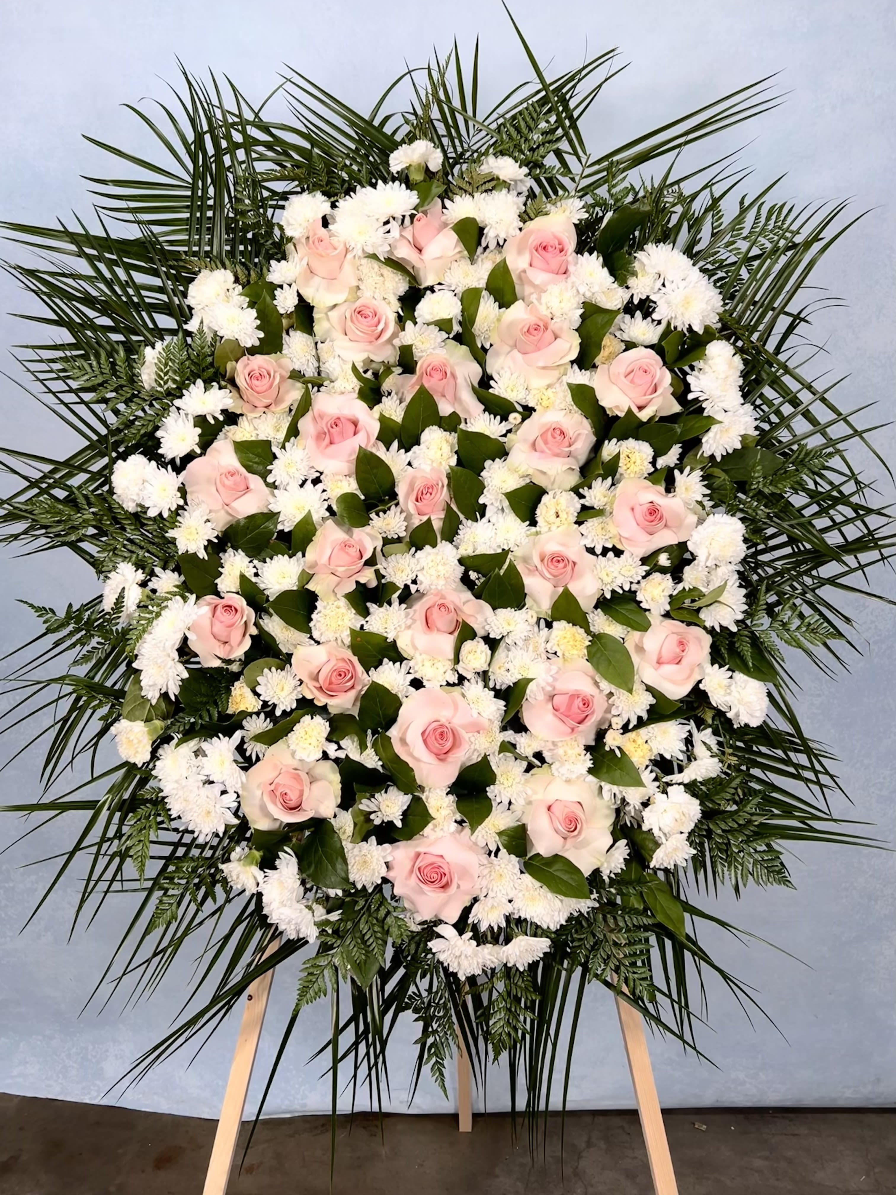 Large standing spray of pink roses and white flowers framed by palm fronds on a wooden easel.