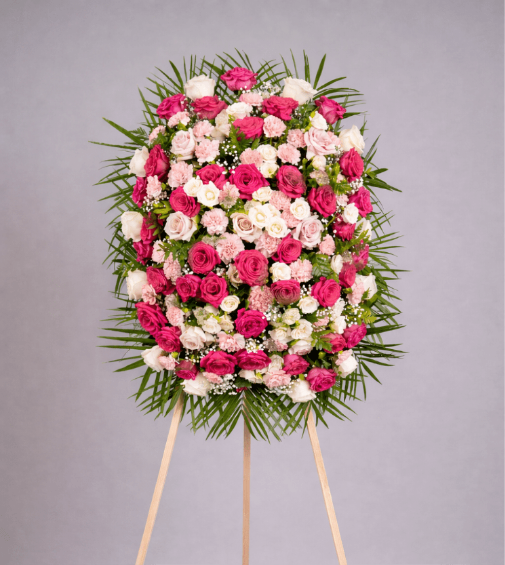 Standing oval spray of pink and white roses and carnations on an easel, framed by a ring of fanned greenery.