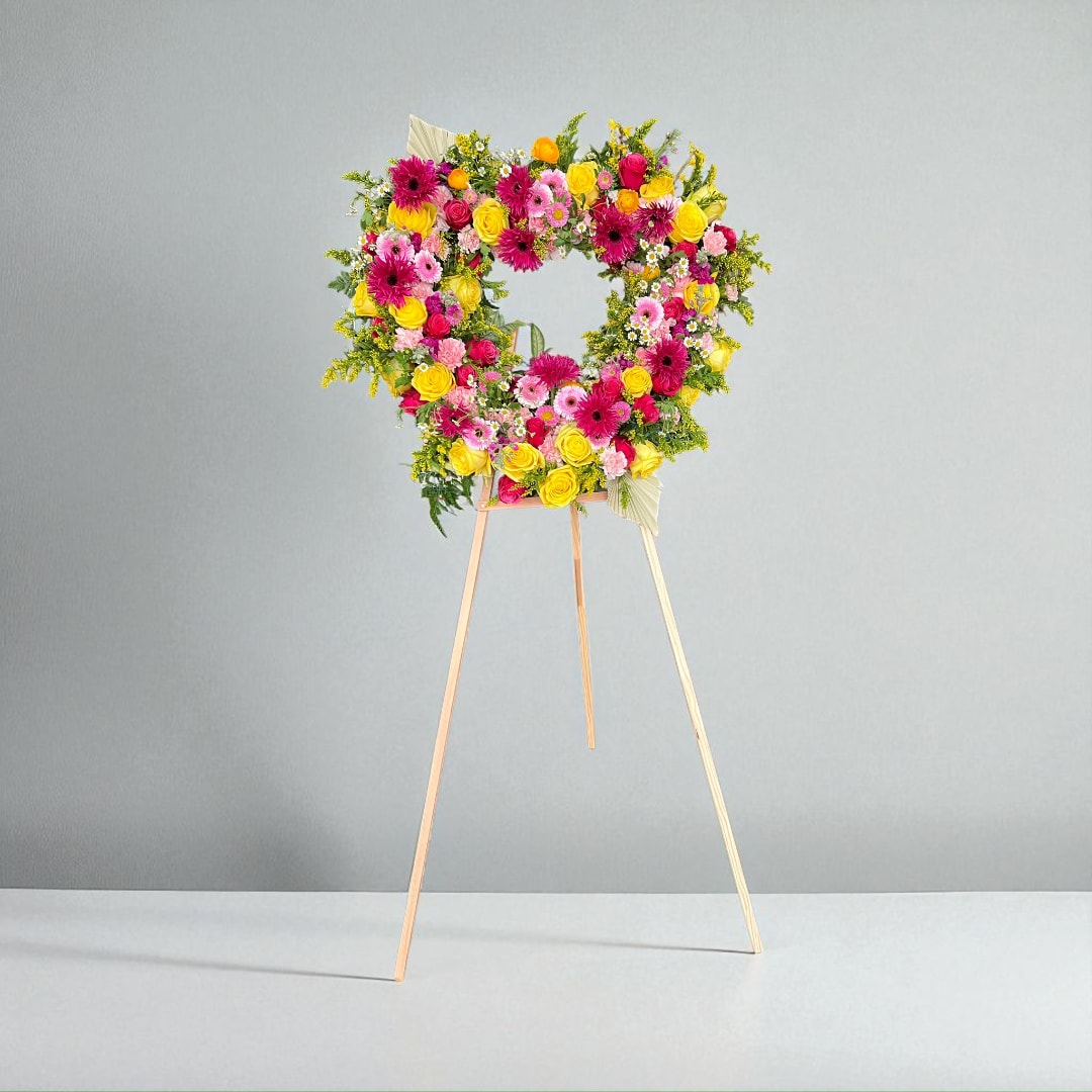 Heart-shaped floral standing wreath on a wooden easel with pink, yellow, and orange blooms
