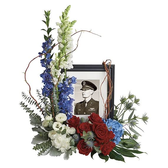 Floral tribute with red roses, blue and white flowers, and a framed military portrait